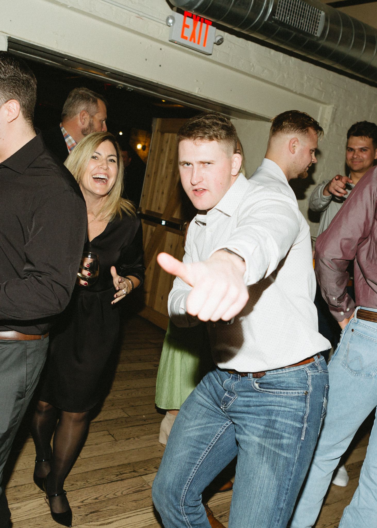 Party guests dancing and laughing at an indoor reception with exposed ceiling.