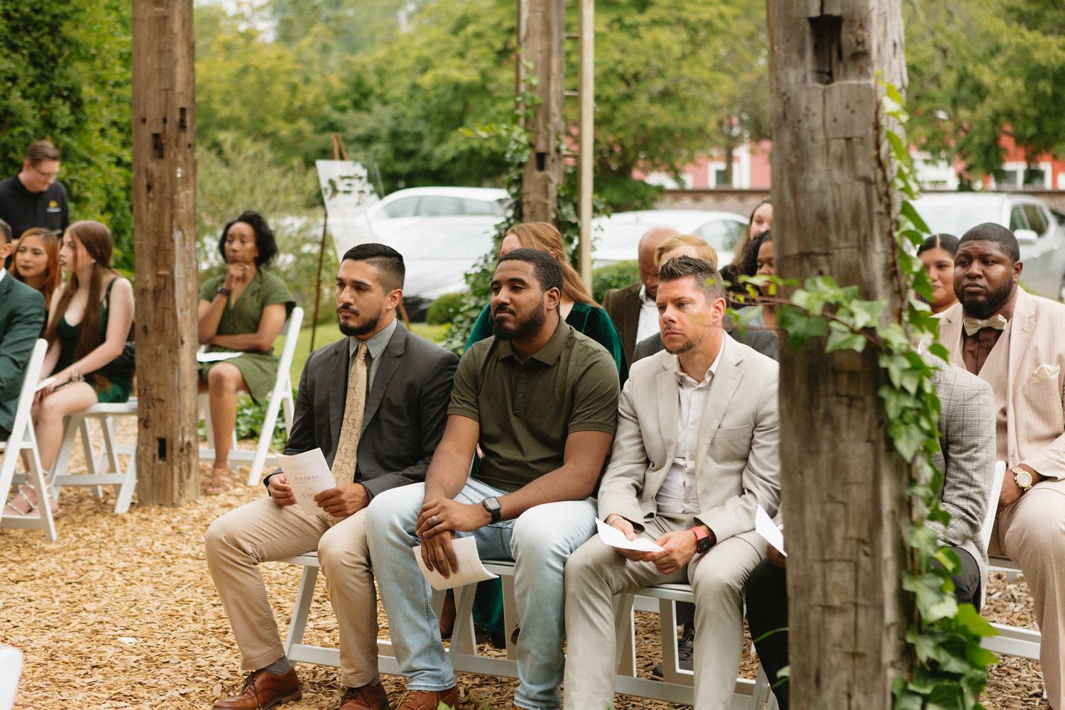 Wedding guests in casual attire seated on wooden chairs in an outdoor garden setting during a ceremony.
