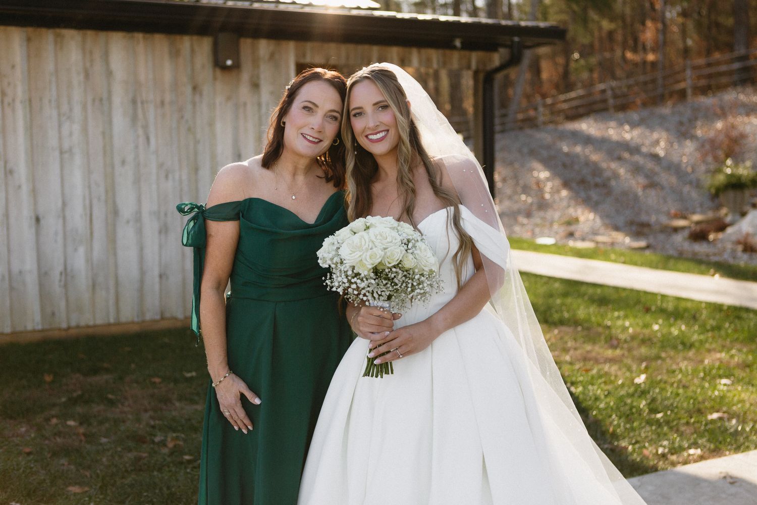 Wedding party with bridesmaids in dark green dresses posing outdoors near wooden barn structure.