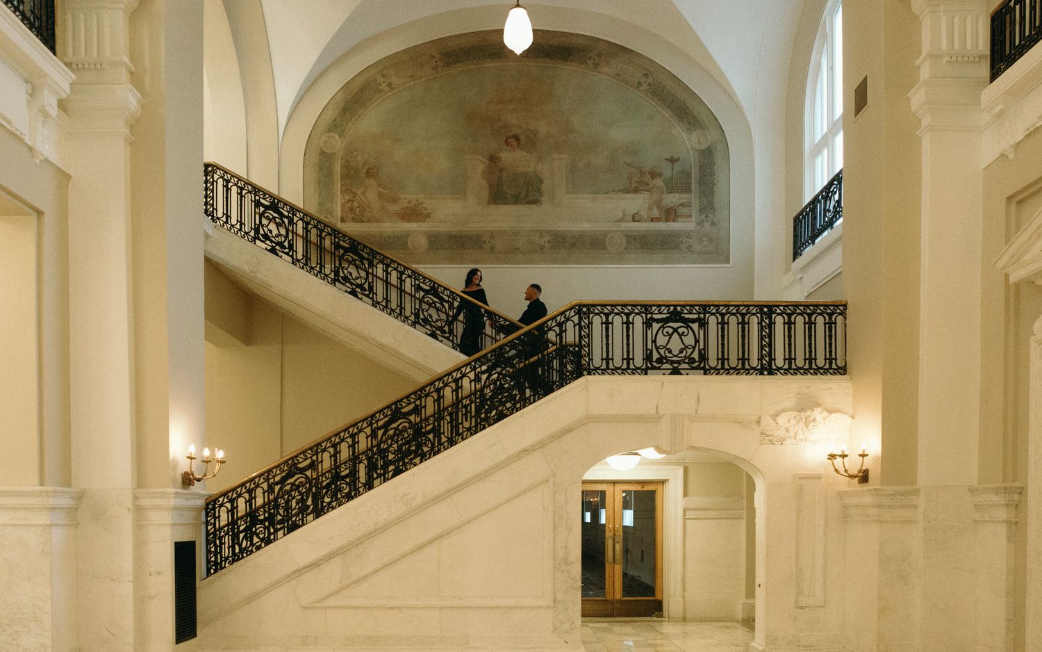 Elegant grand staircase with ornate black iron railings and cream walls inside a classical building interior.