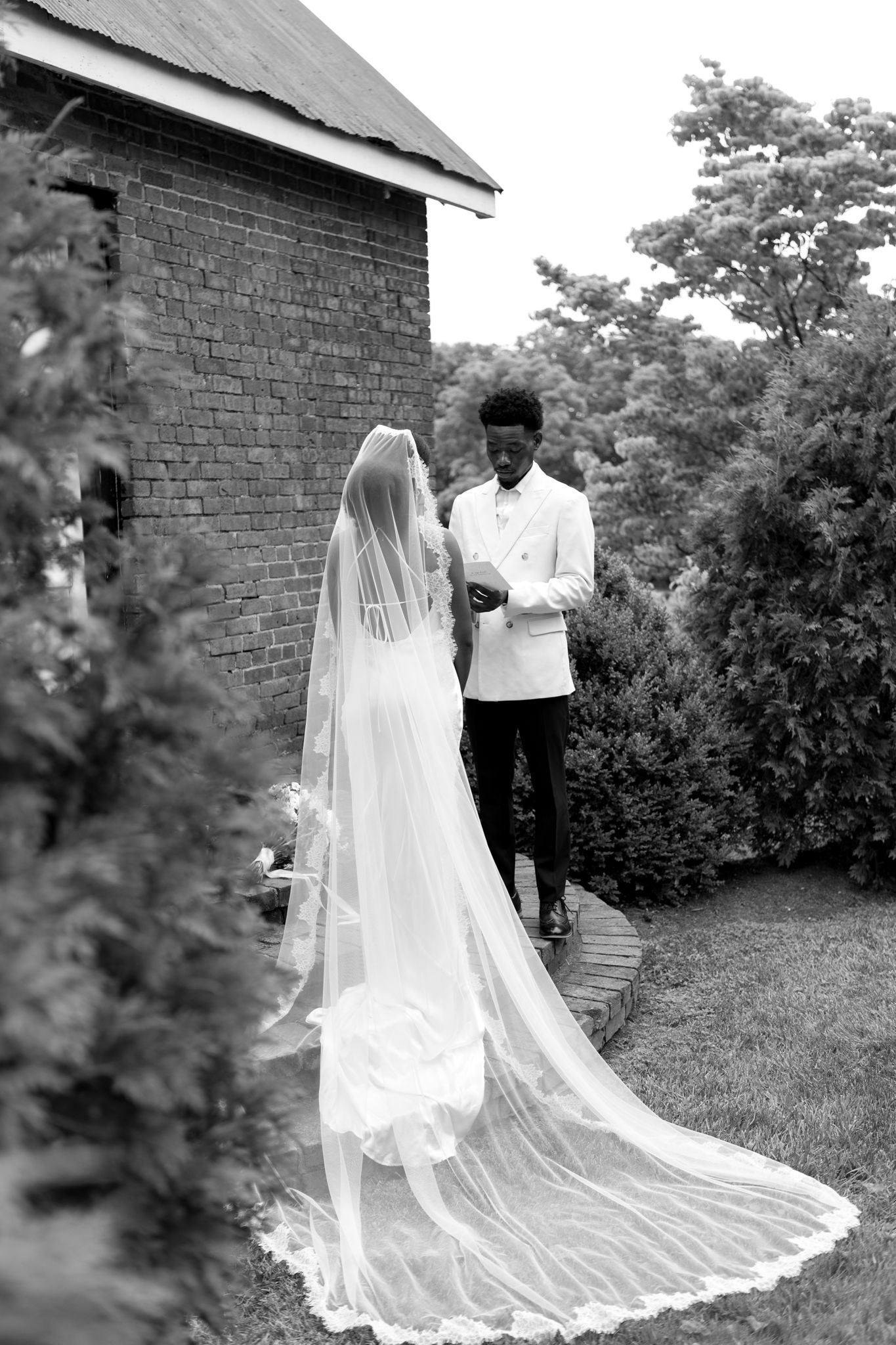 Elegant black and white wedding portrait with long trailing veil against rustic brick building.
