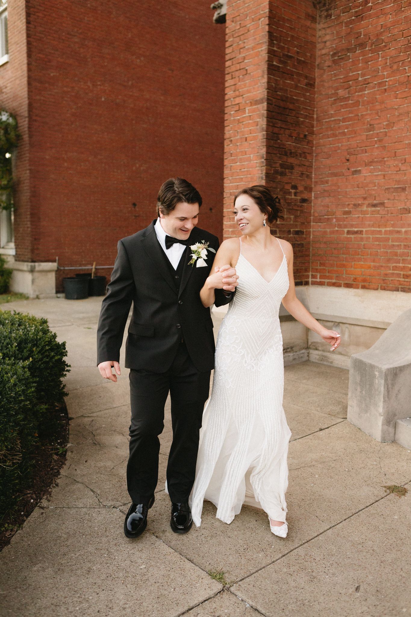 A newlywed couple walking and laughing together outside a red brick building on their wedding day.