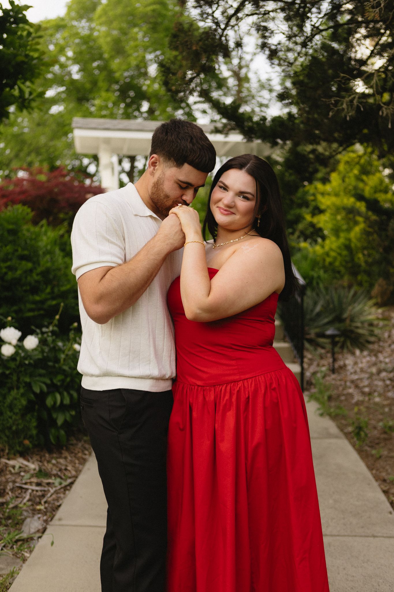 Couple embraces on garden pathway as woman in flowing red dress smiles joyfully.