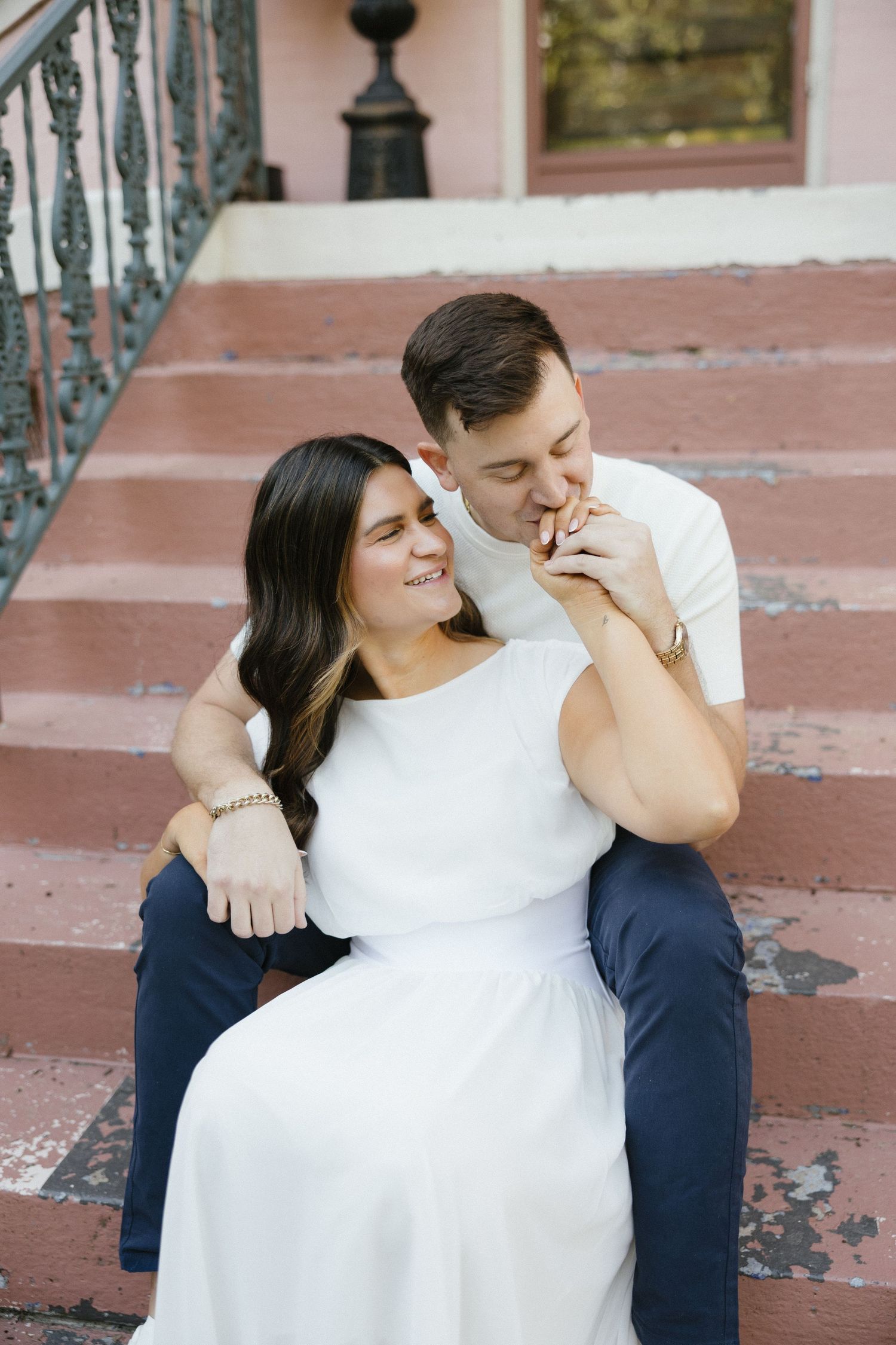 Couple sitting on pink steps, man kissing woman's hand while she smiles, wearing white dress and navy pants.