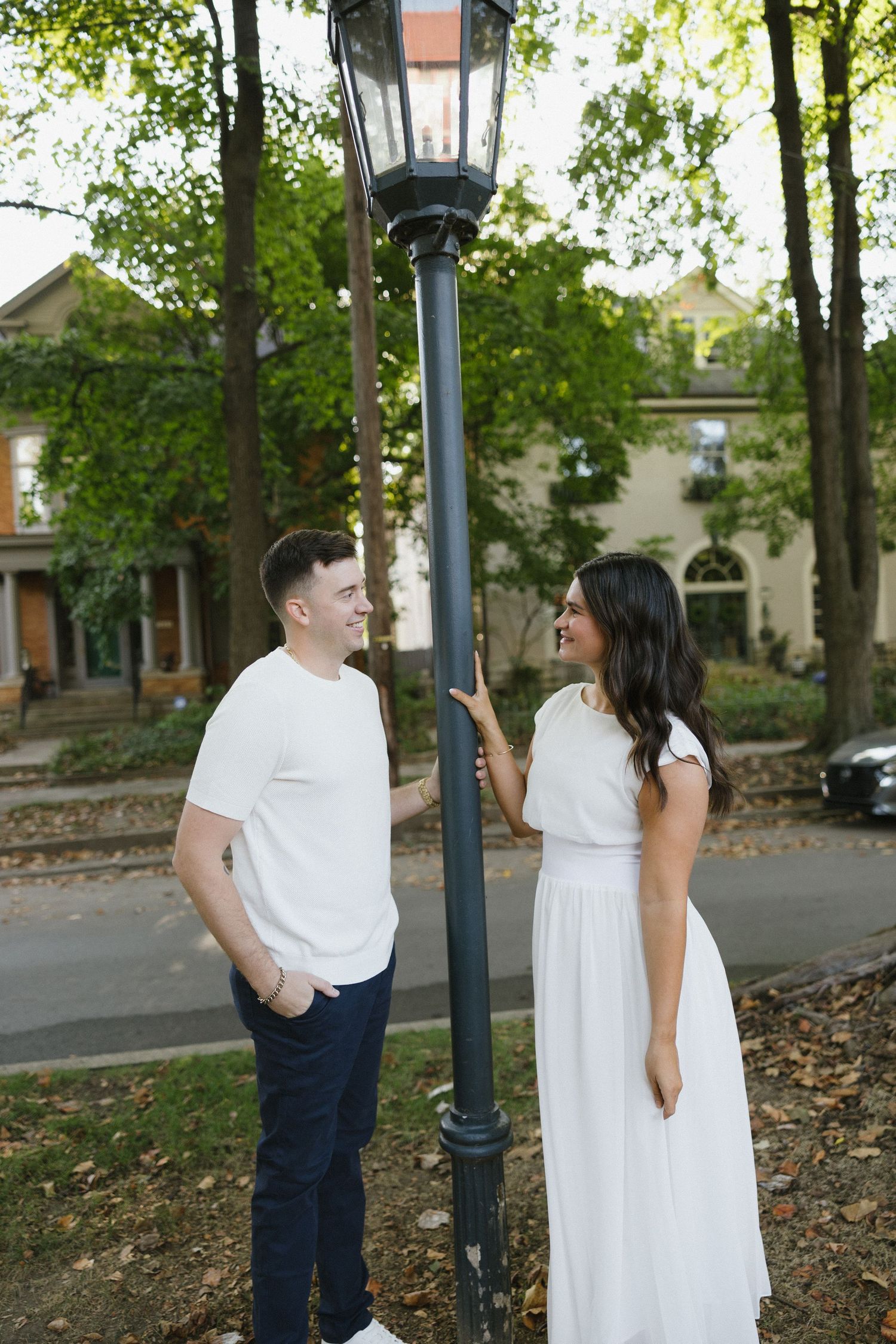 A couple in white outfits stands by a vintage street lamp in a tree-lined neighborhood, smiling at each other.