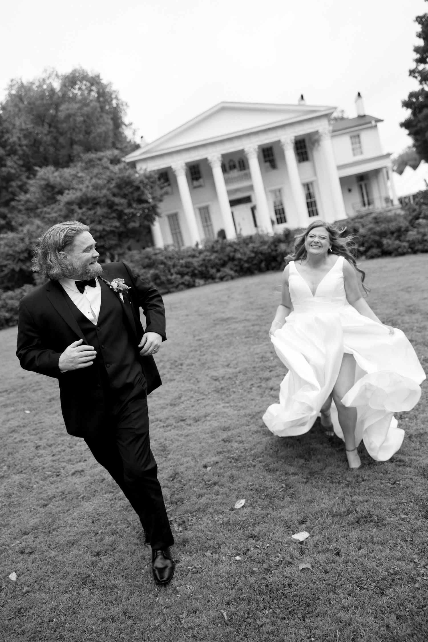 Black and white photo of a laughing bride and groom running across a lawn in front of a grand white plantation-style mansion.