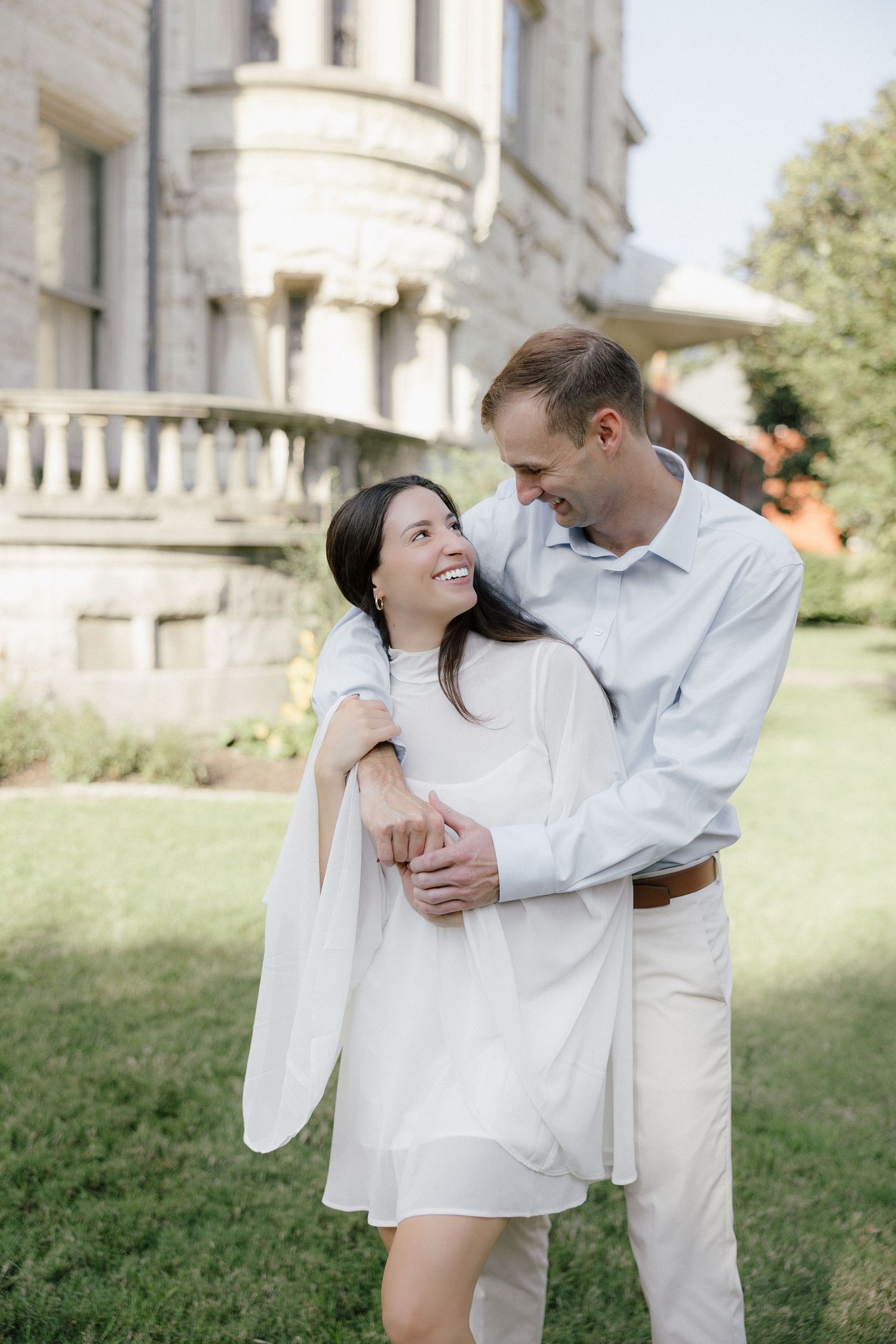 Couple embracing and smiling at each other on green lawn in front of a classic stone building.