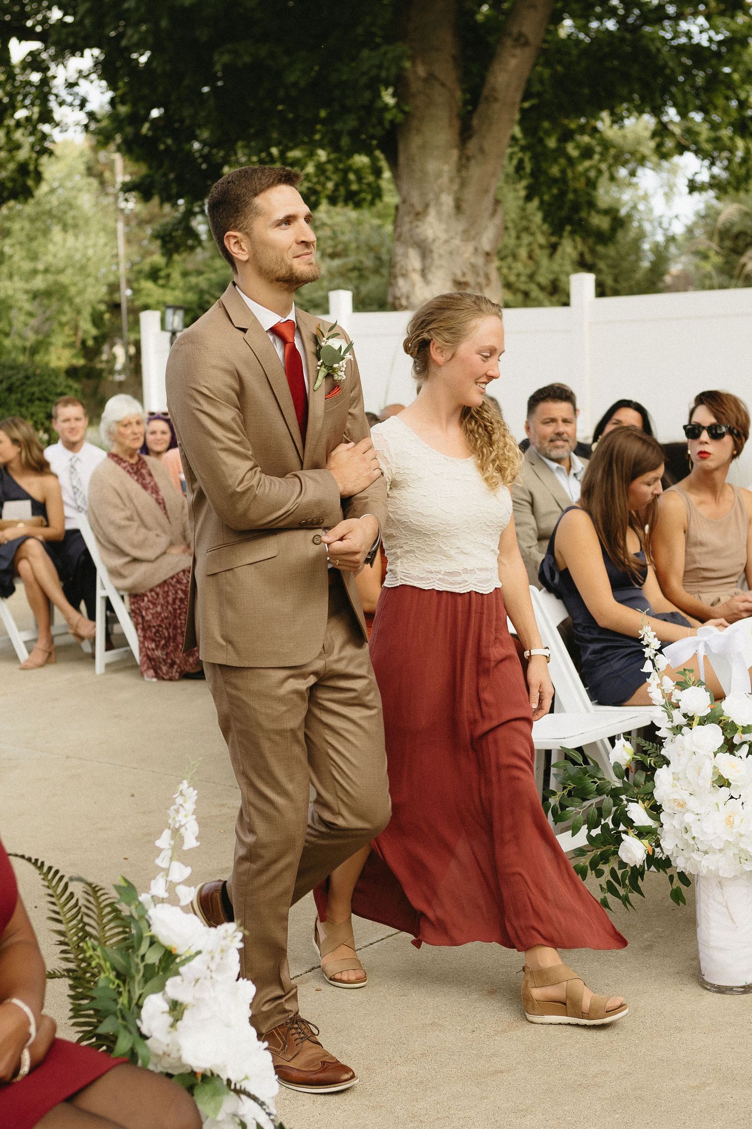 Wedding party walking outdoors in burgundy and tan attire with white floral decorations.
