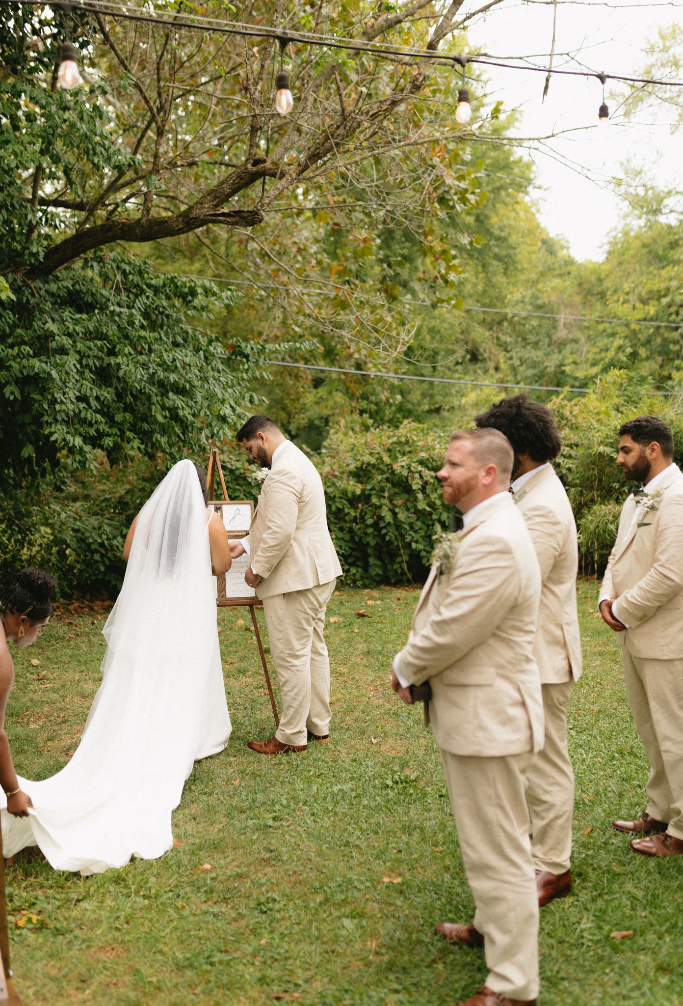 A wedding ceremony takes place outdoors with people in beige suits standing in a garden setting beneath string lights.