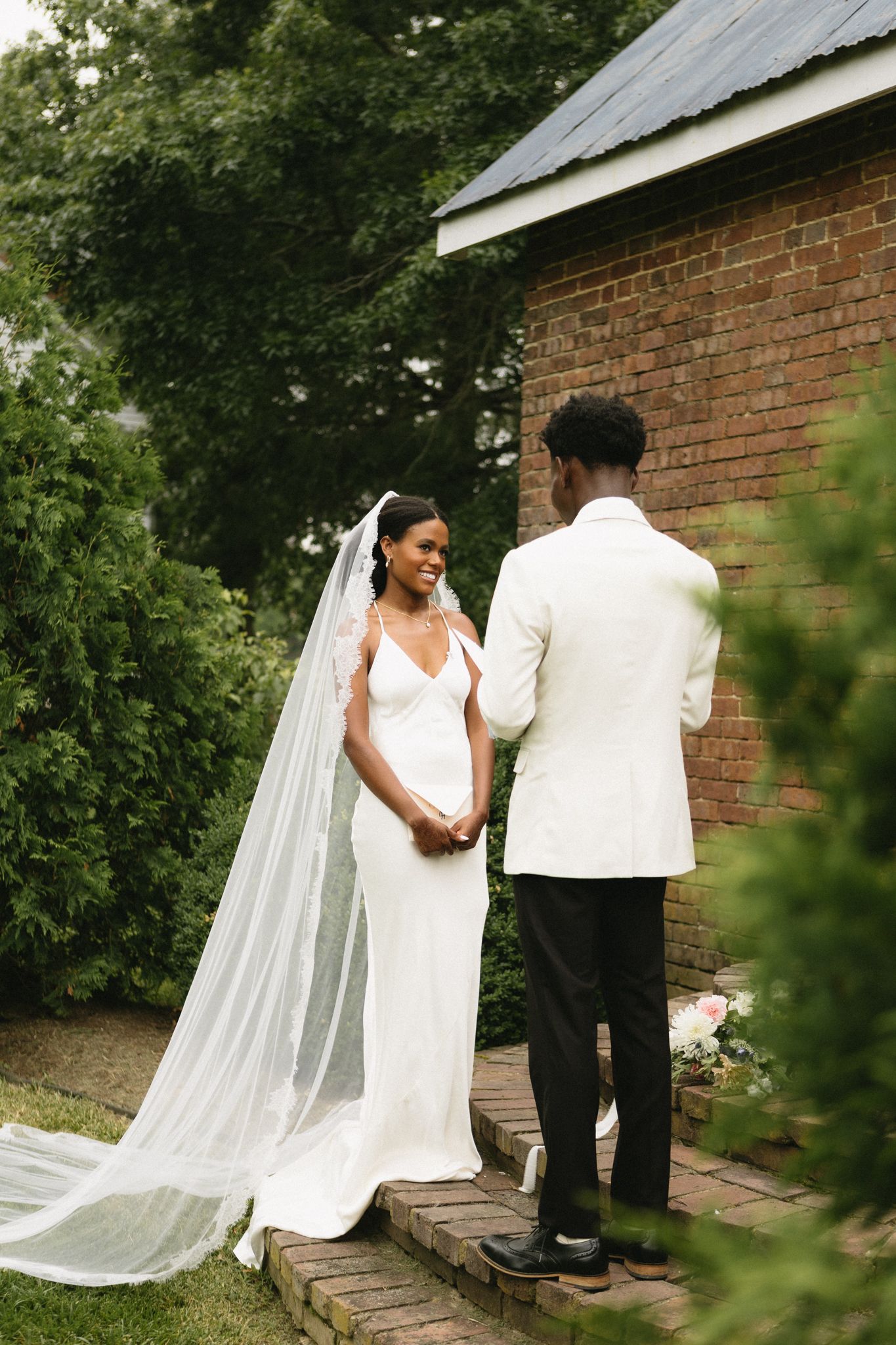 Outdoor wedding couple poses against brick wall with flowing white veil in garden setting.