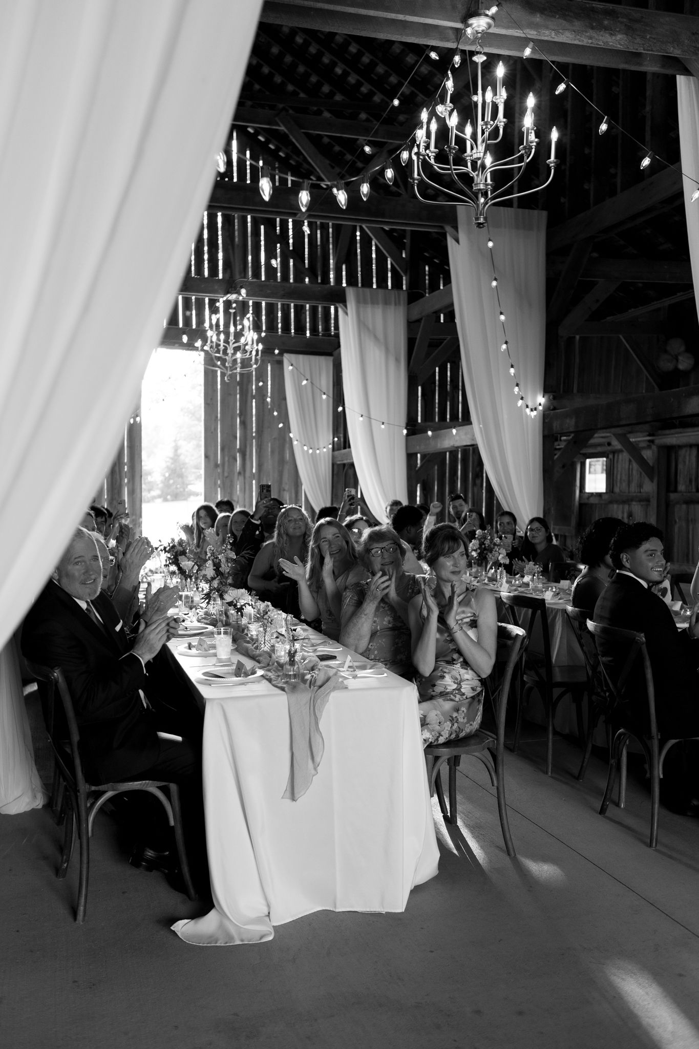Wedding reception in a rustic barn venue with elegant chandeliers and white draping.
