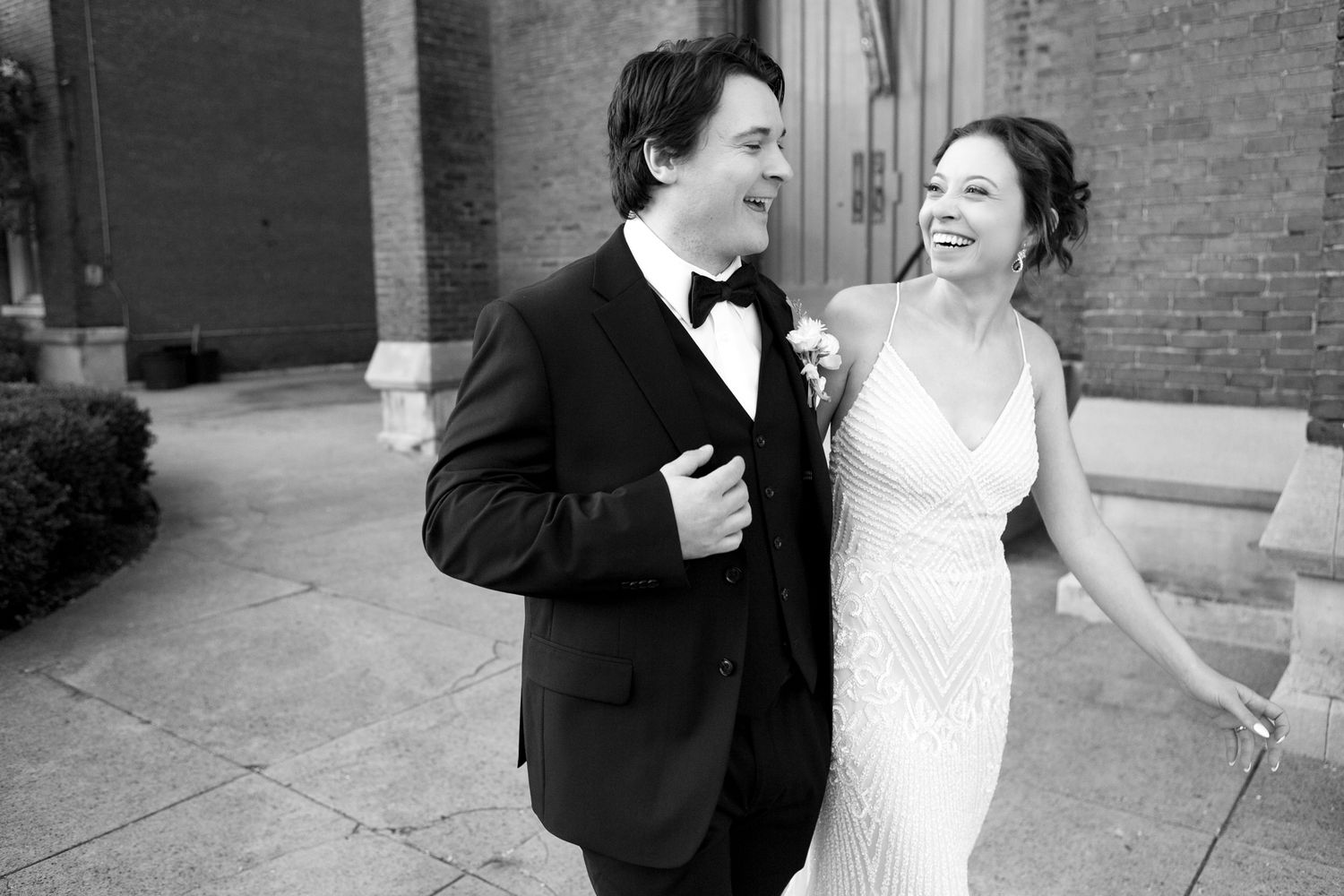 An elegant couple sharing a joyful moment outside a brick building in formal attire.
