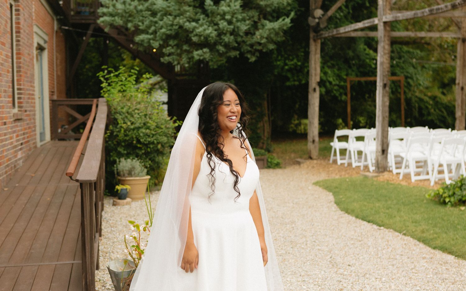 A bride in a flowing white wedding dress and long veil stands outdoors near a wooden deck and garden ceremony setup.