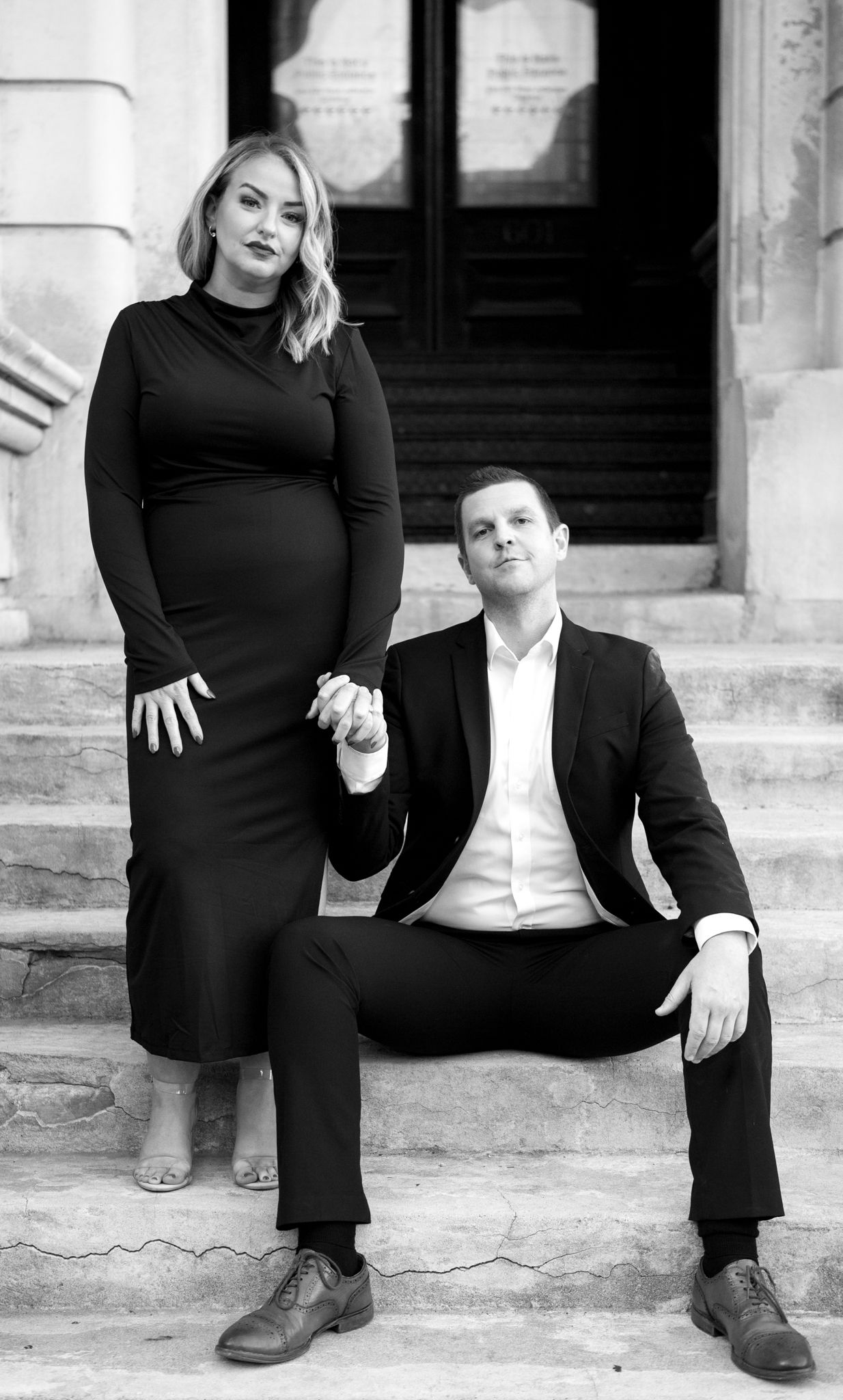 A formal couple poses on stone steps in black and white, creating an elegant engagement portrait.