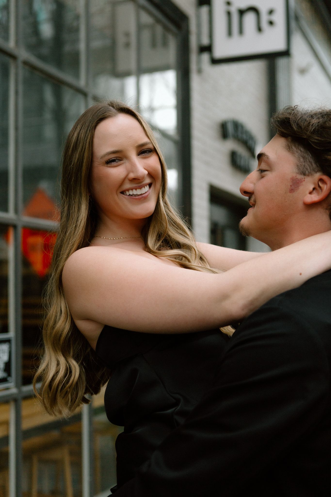 Engaged couple smiling and embracing, with the woman showing her engagement ring, captured in downtown Louisville