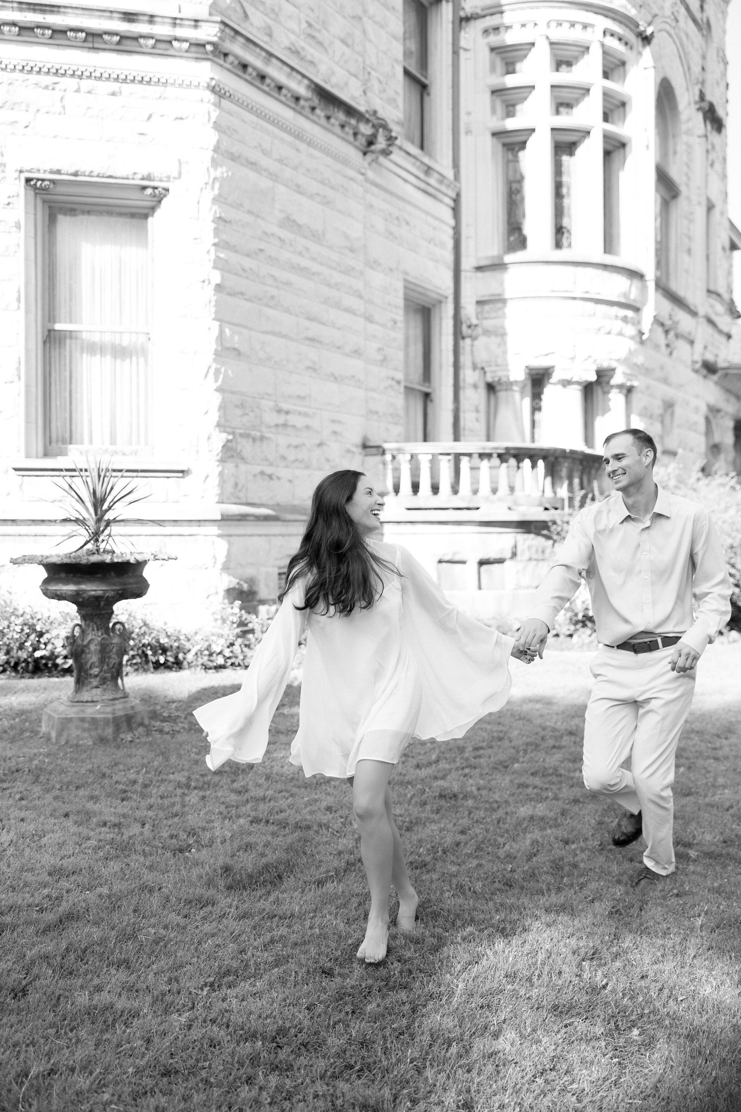 Black and white photo of a couple holding hands and walking on grass in front of an elegant historic building.