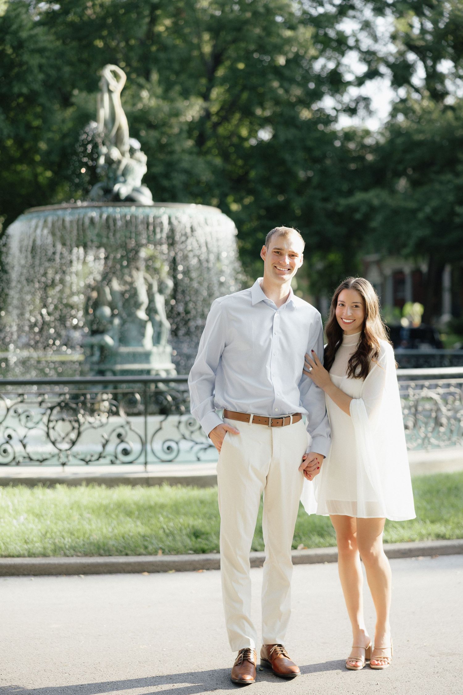 Smiling couple posing in front of an ornate fountain in a lush green park, woman in white dress holding mans hand.