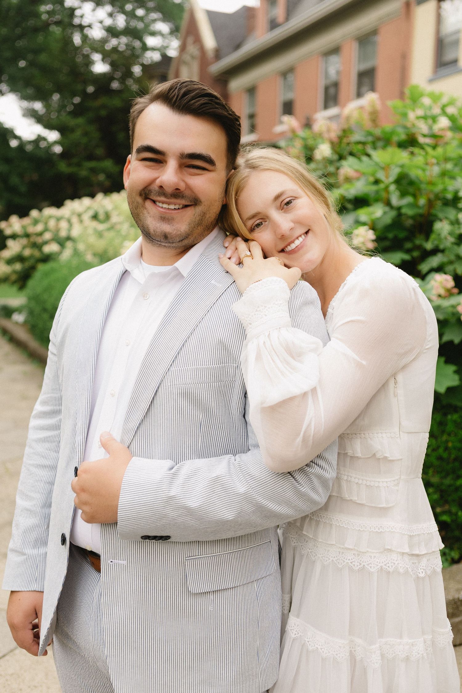 Happy couple posing together outdoors, woman in white dress leaning on mans shoulder in gray suit.