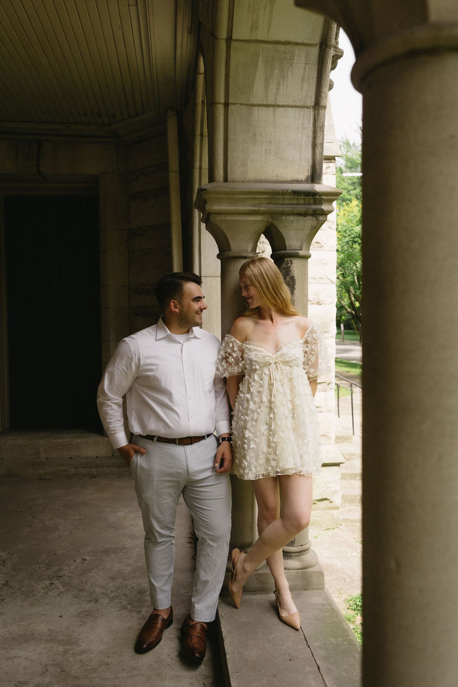 Engaged couple smiles at each other while standing among stone columns during elegant outdoor engagement session.