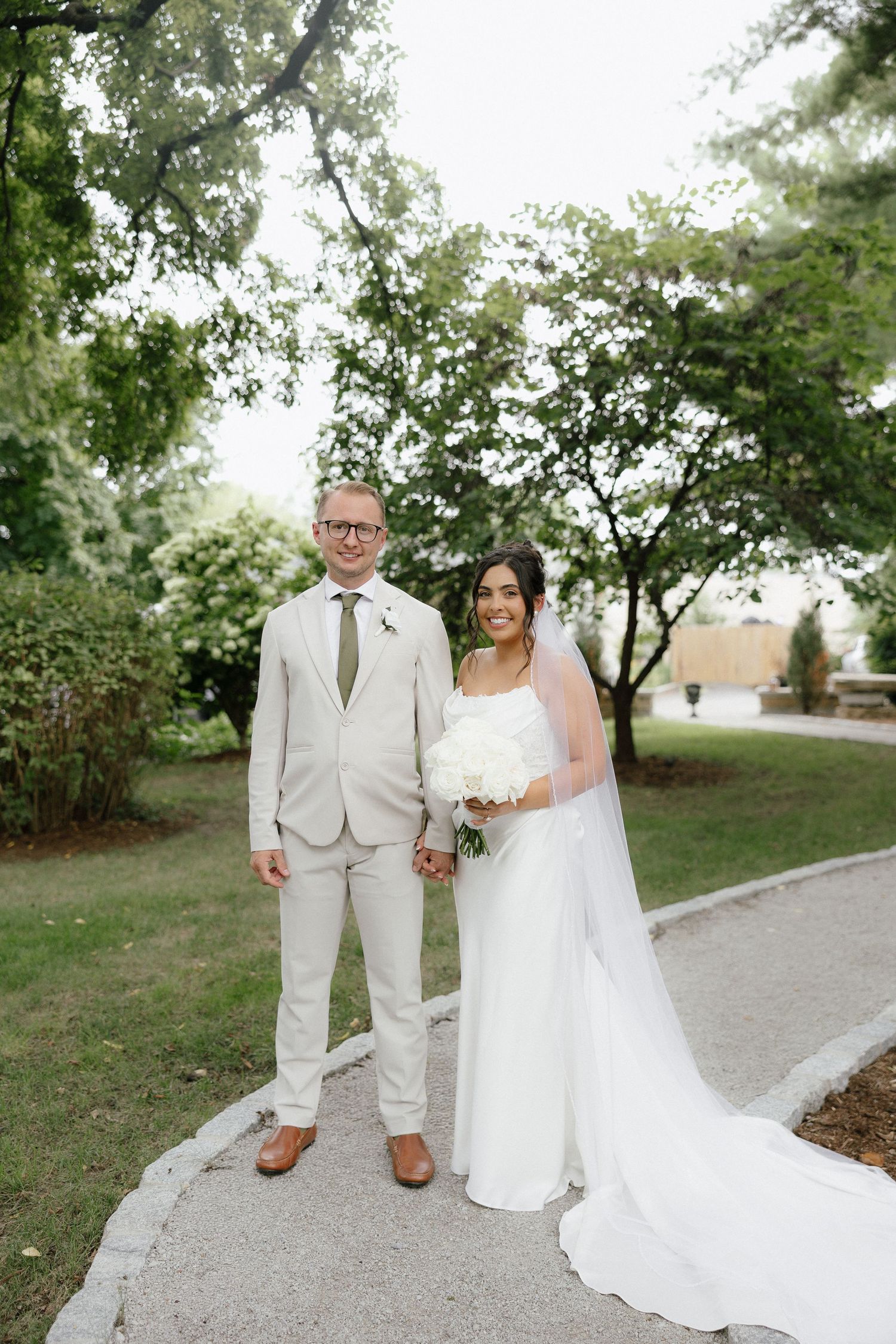 Bride and groom holding hands on garden path, bride in white gown with veil, groom in beige suit.