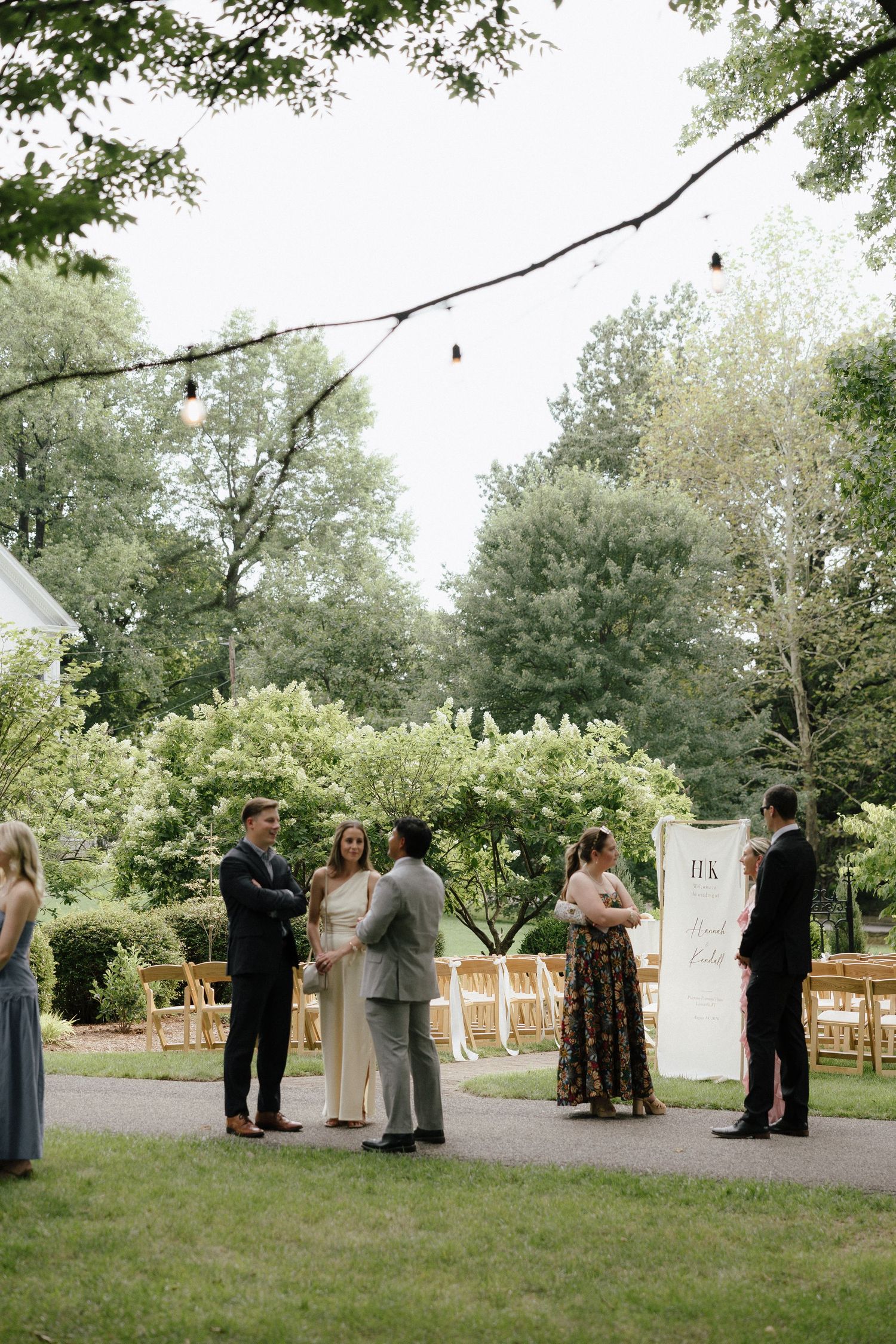 Wedding guests mingling outdoors under string lights near a monogram sign in a lush garden setting.