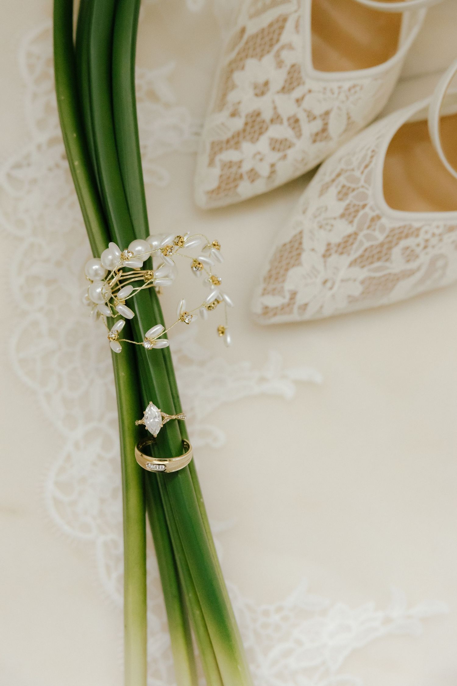 Bridal flat shoes with lace detailing beside wedding rings on green stems with pearl accents.
