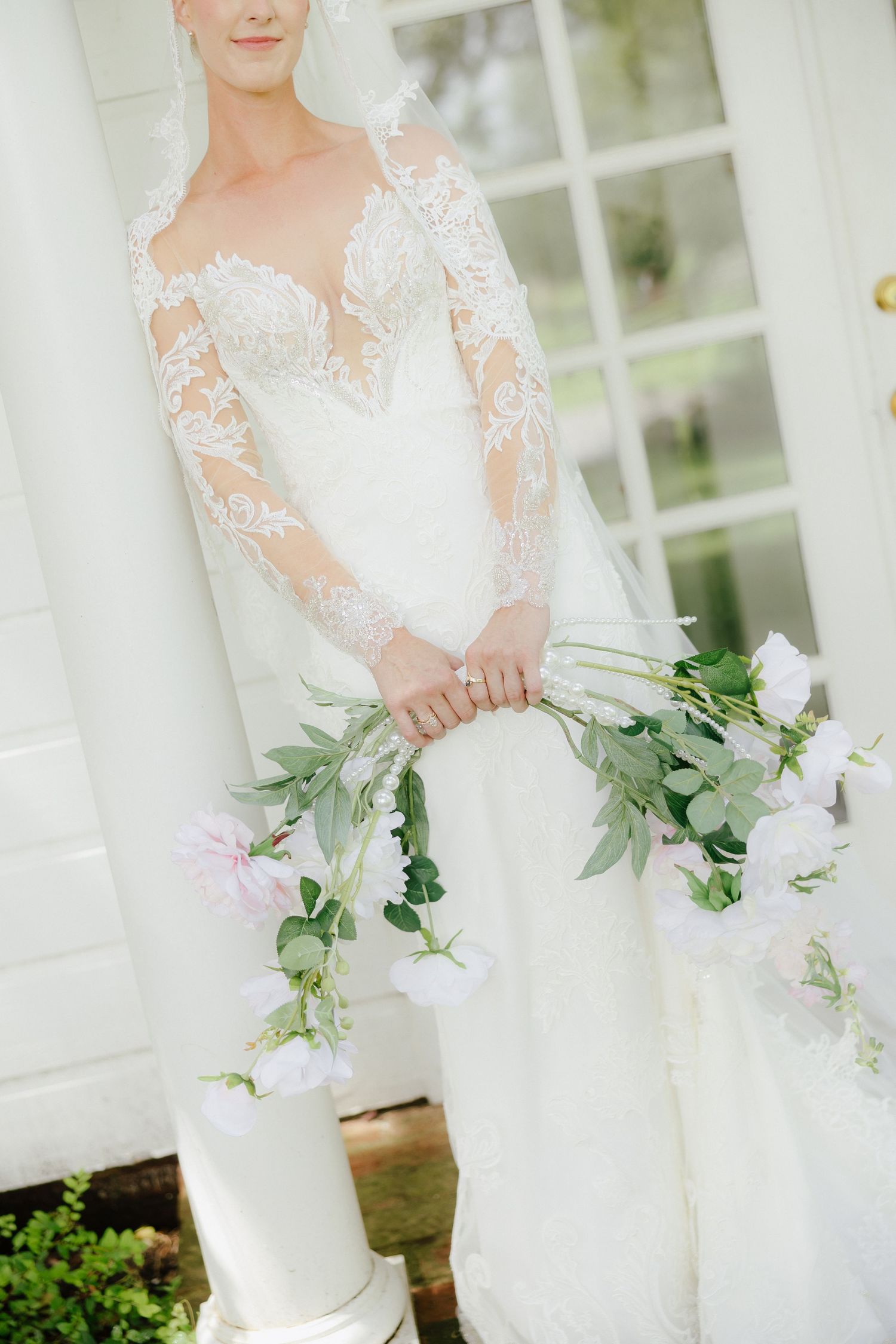 Bride in long-sleeve lace wedding gown holds cascading pink floral bouquet near white French doors.