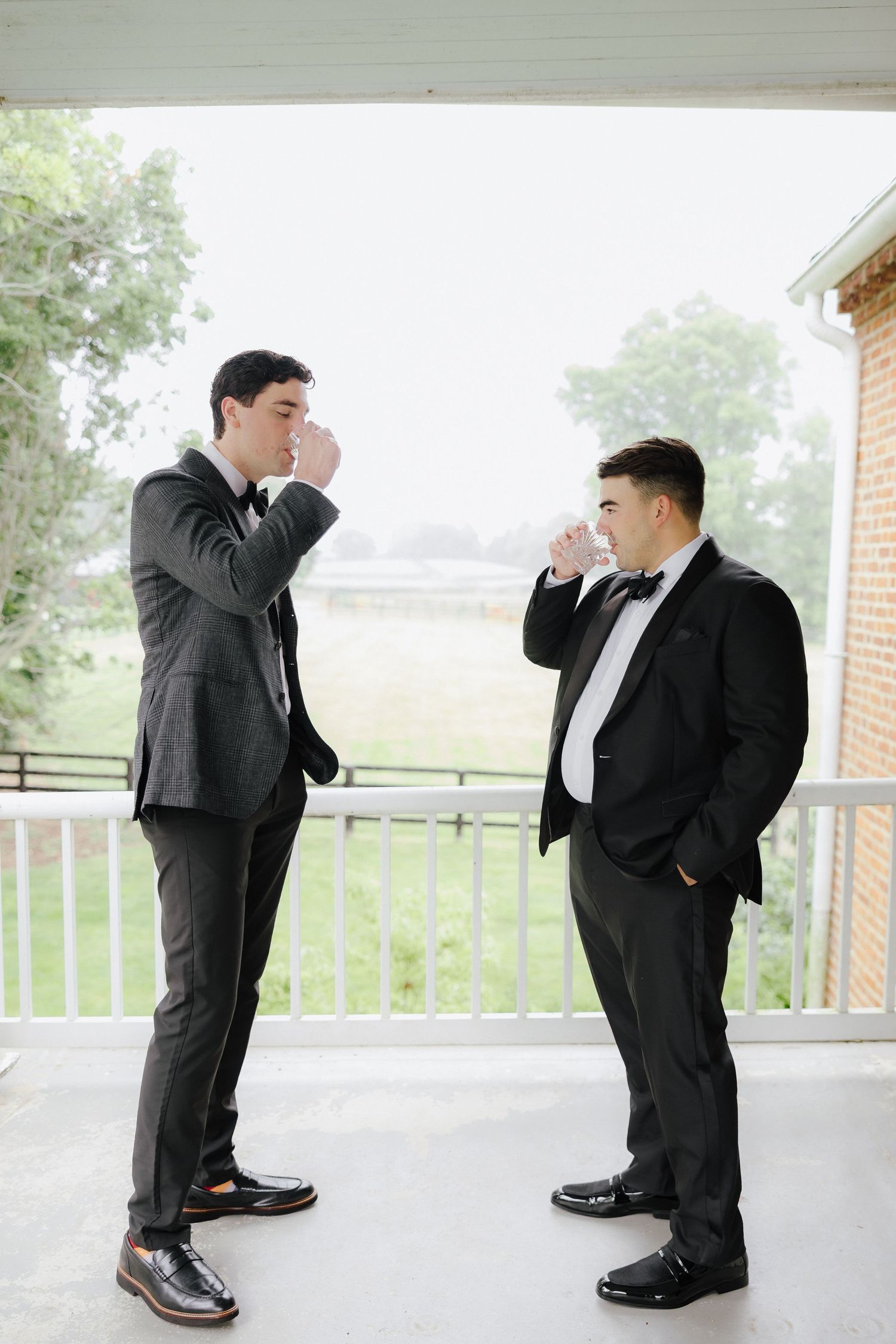 Two groomsmen in black suits share a toast on an outdoor porch before a wedding ceremony.
