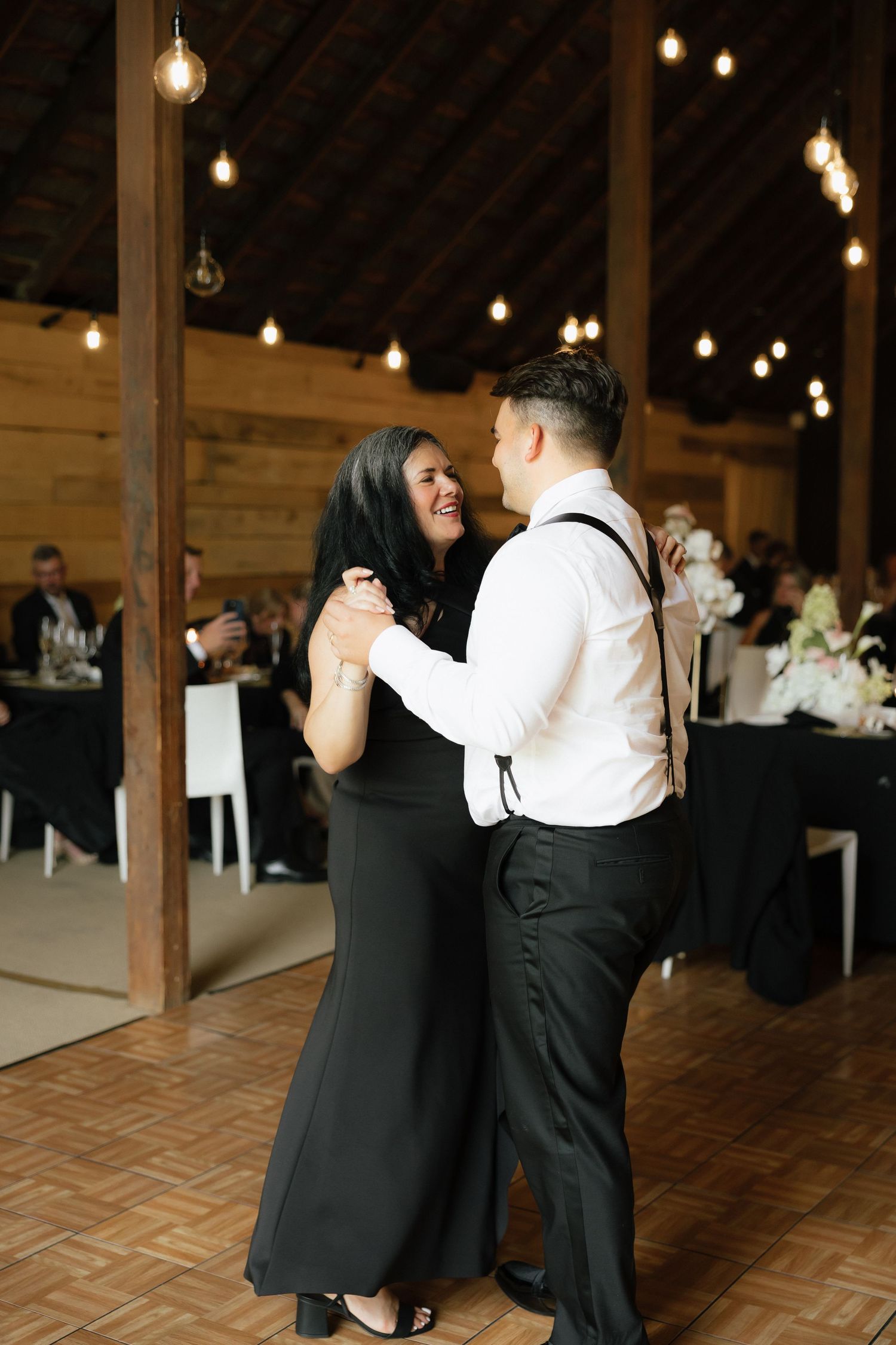 A couple shares a mother-son dance in a warmly lit barn venue during a wedding reception.