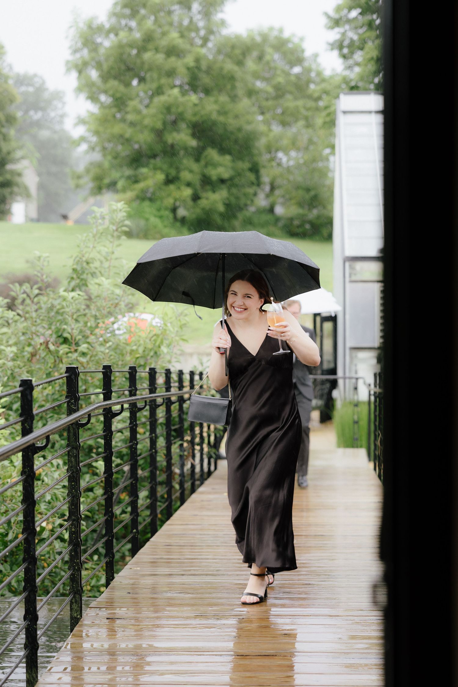 Woman in black satin dress walking with umbrella and cocktail in the rain at outdoor wedding venue.