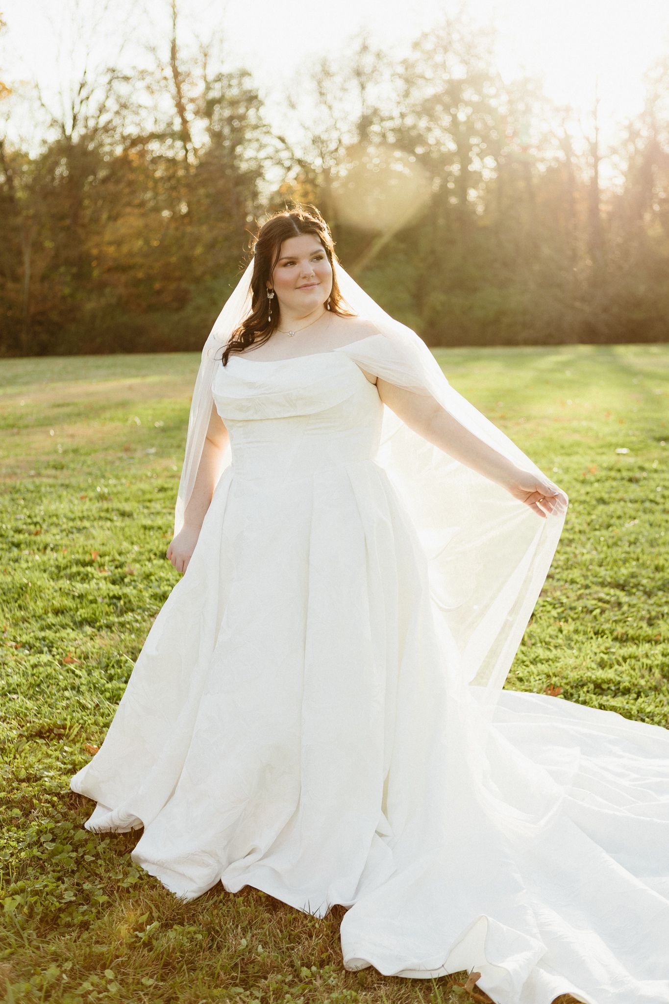 A flowing white wedding dress billows in the golden sunlight on a grassy field during sunset.