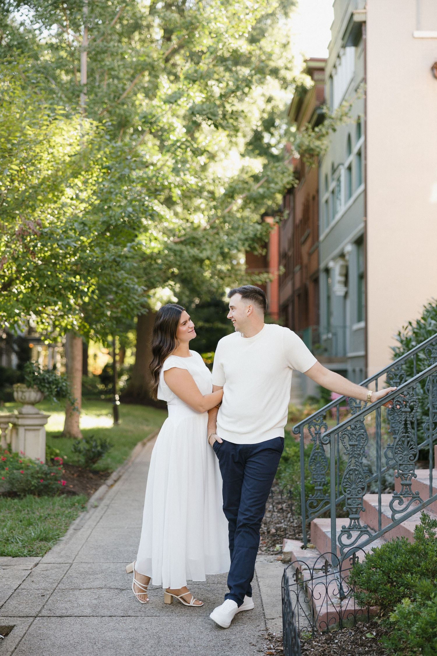Couple in white outfits walk hand-in-hand on a tree-lined sidewalk beside colorful townhomes.