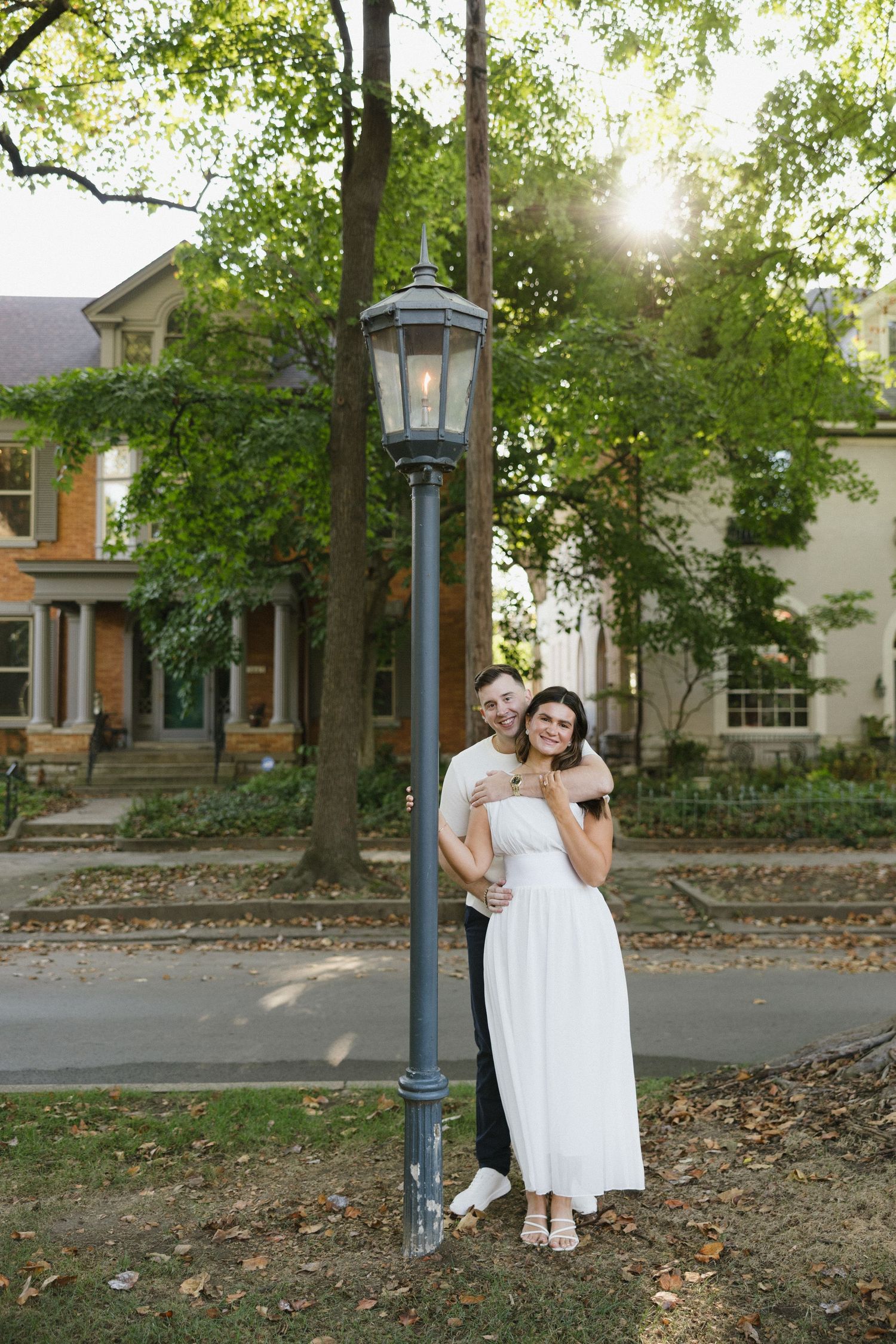 Romantic couple embracing by vintage street lamp in tree-lined neighborhood at golden hour.