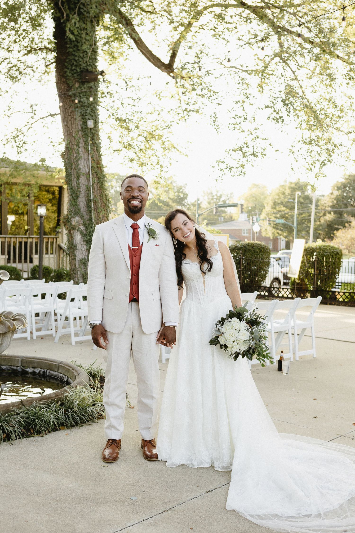 Wedding couple walks together on an outdoor pathway surrounded by trees and white chairs.