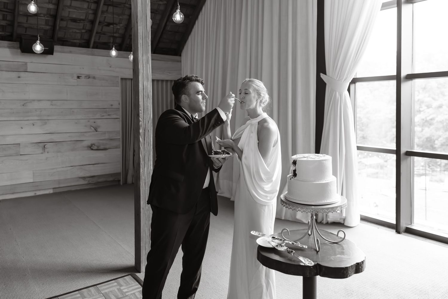 Black and white photo of groom feeding bride wedding cake at elegant barn venue.