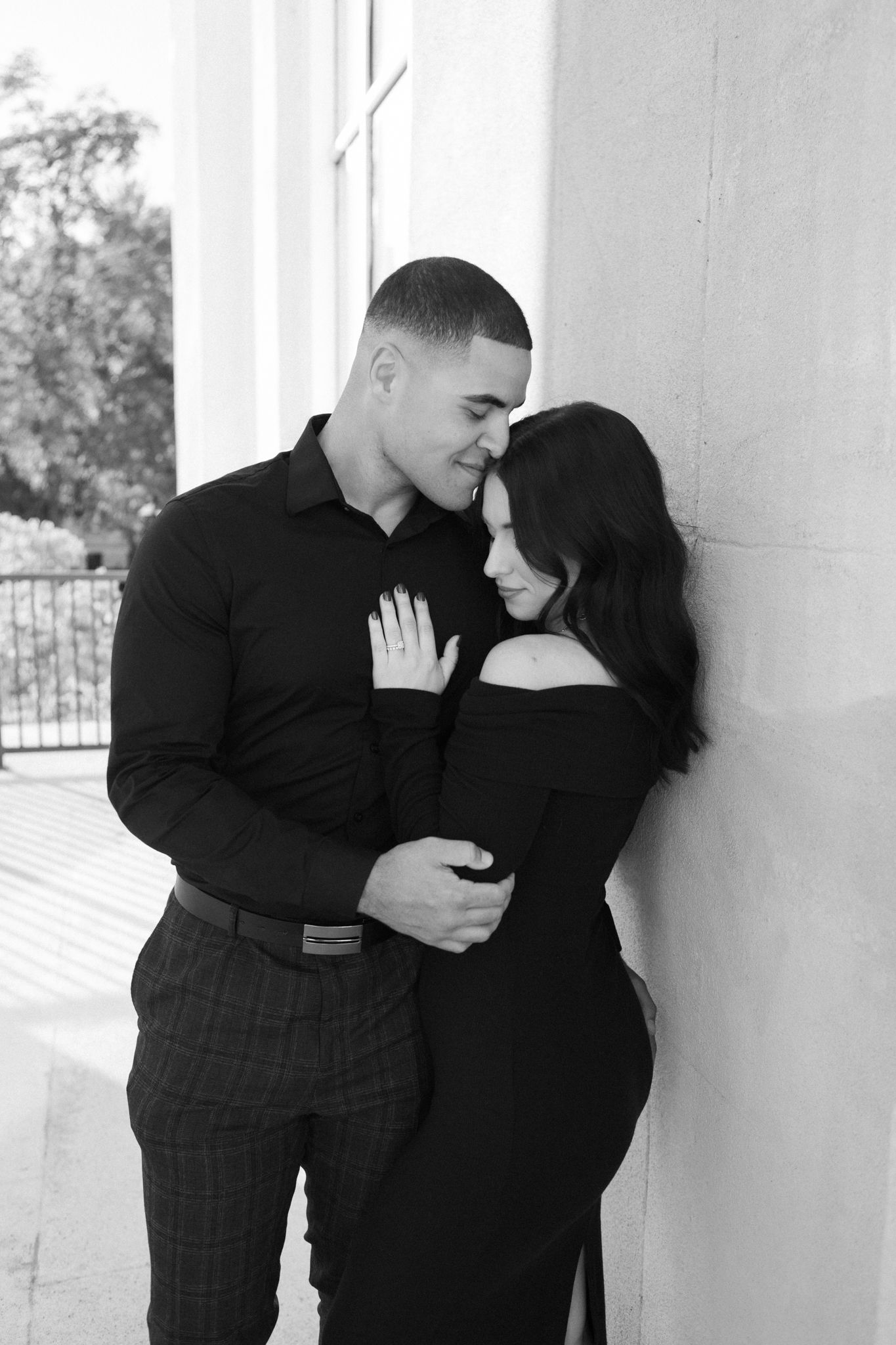 A romantic black and white portrait of a couple embracing against a white wall on a porch.