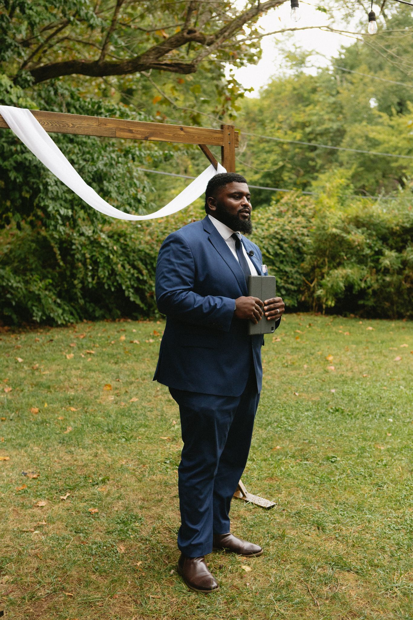 A person in a navy blue suit stands in a garden setting beneath white wedding decorations on a fall day.