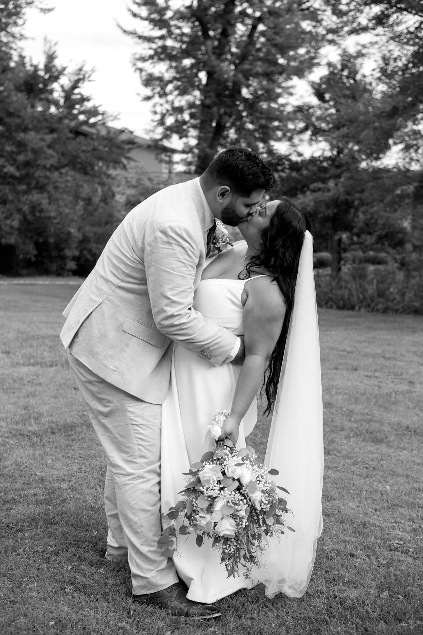 A romantic black and white wedding photo shows a couple sharing a kiss outdoors with a floral bouquet in hand.