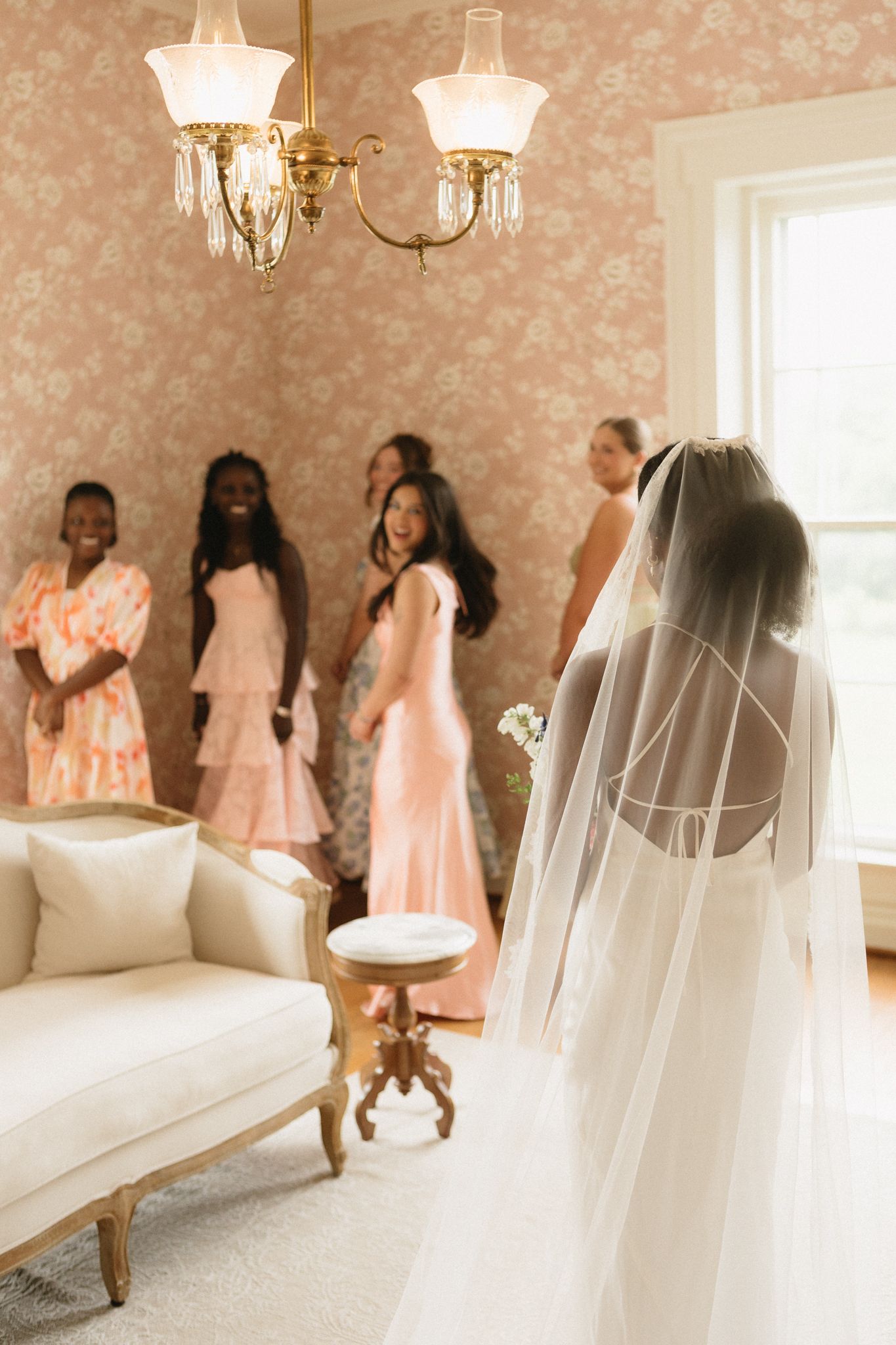 Bridesmaids in soft pink dresses gather in an elegantly decorated room with chandelier and floral wallpaper.
