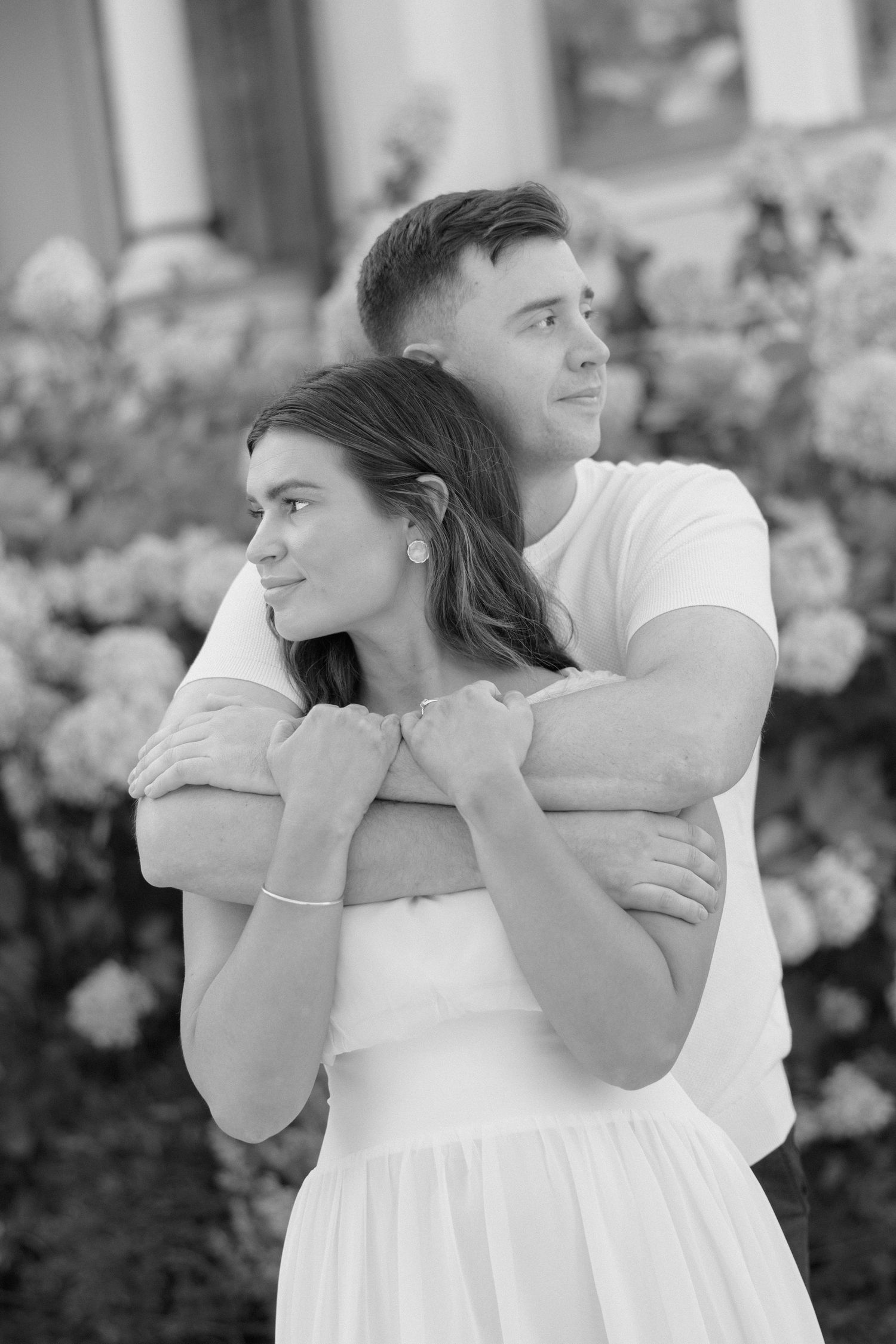 Black and white photo of a couple embracing in front of flowering bushes, man hugging woman from behind.