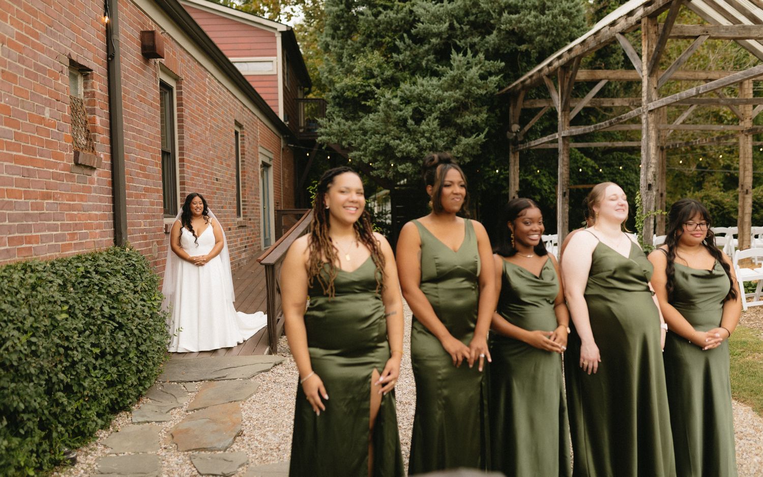 A group of bridesmaids in matching olive green dresses laugh together outside a brick building.