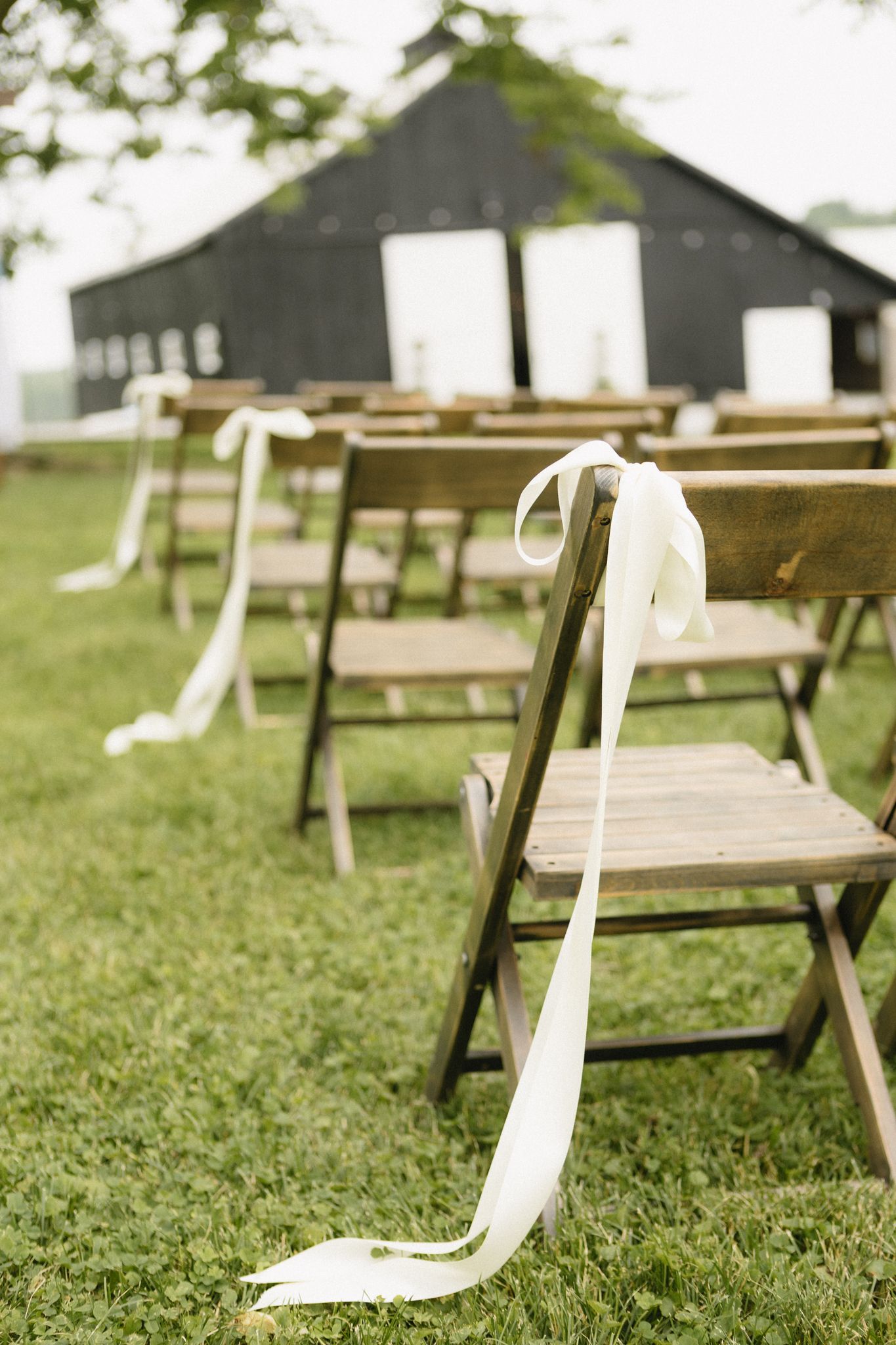 Rustic wooden picnic tables arranged on a grassy lawn outside dark barns during a daytime outdoor event.