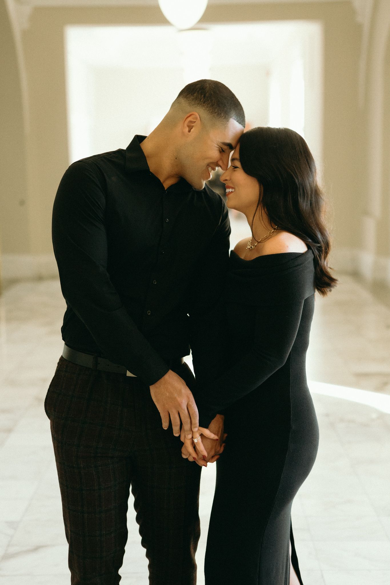 A romantic couple dressed in black share an intimate moment in a softly lit hallway.