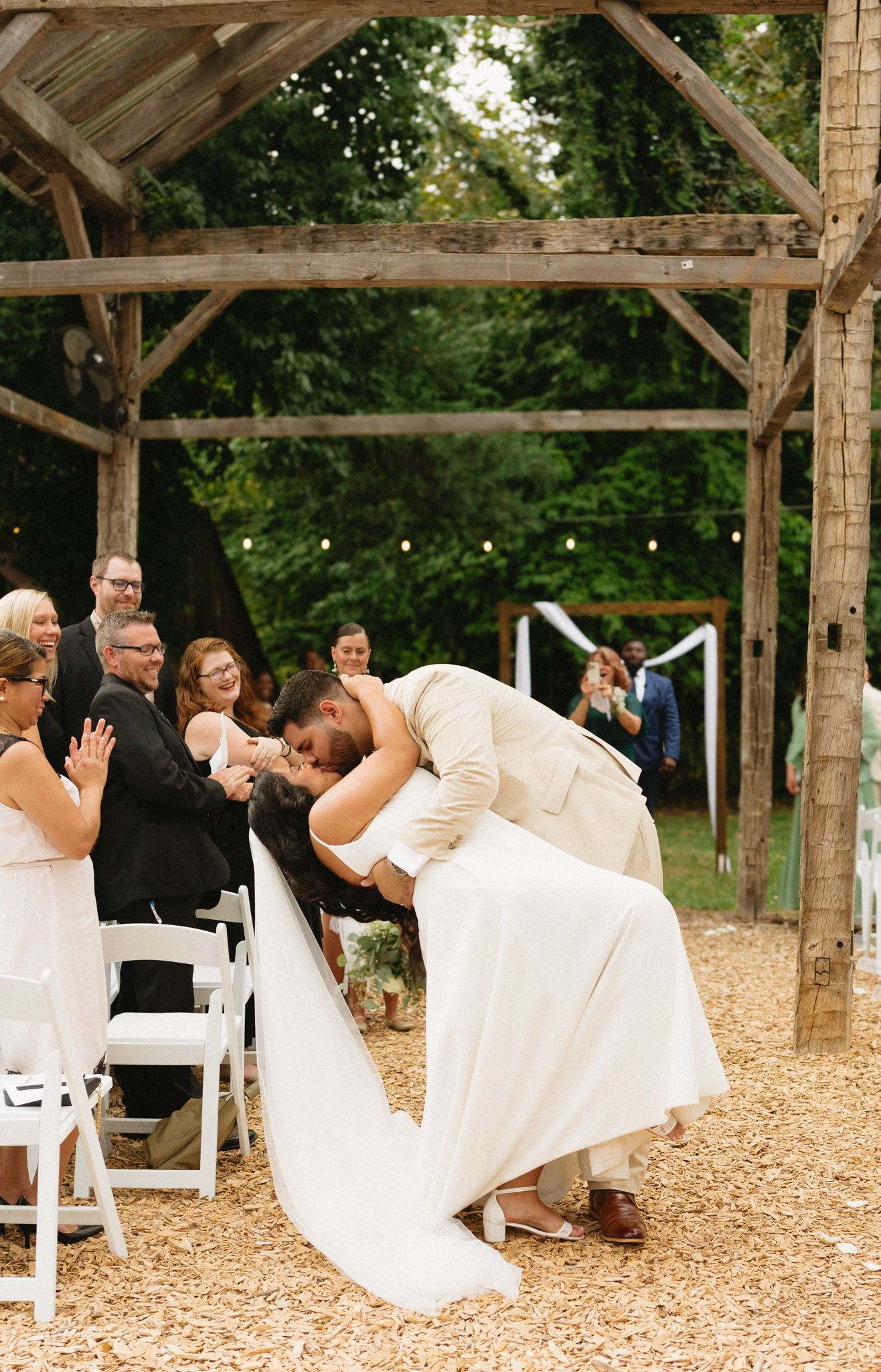 A sweet moment during a rustic outdoor wedding ceremony beneath a wooden beam structure on mulch-covered ground.