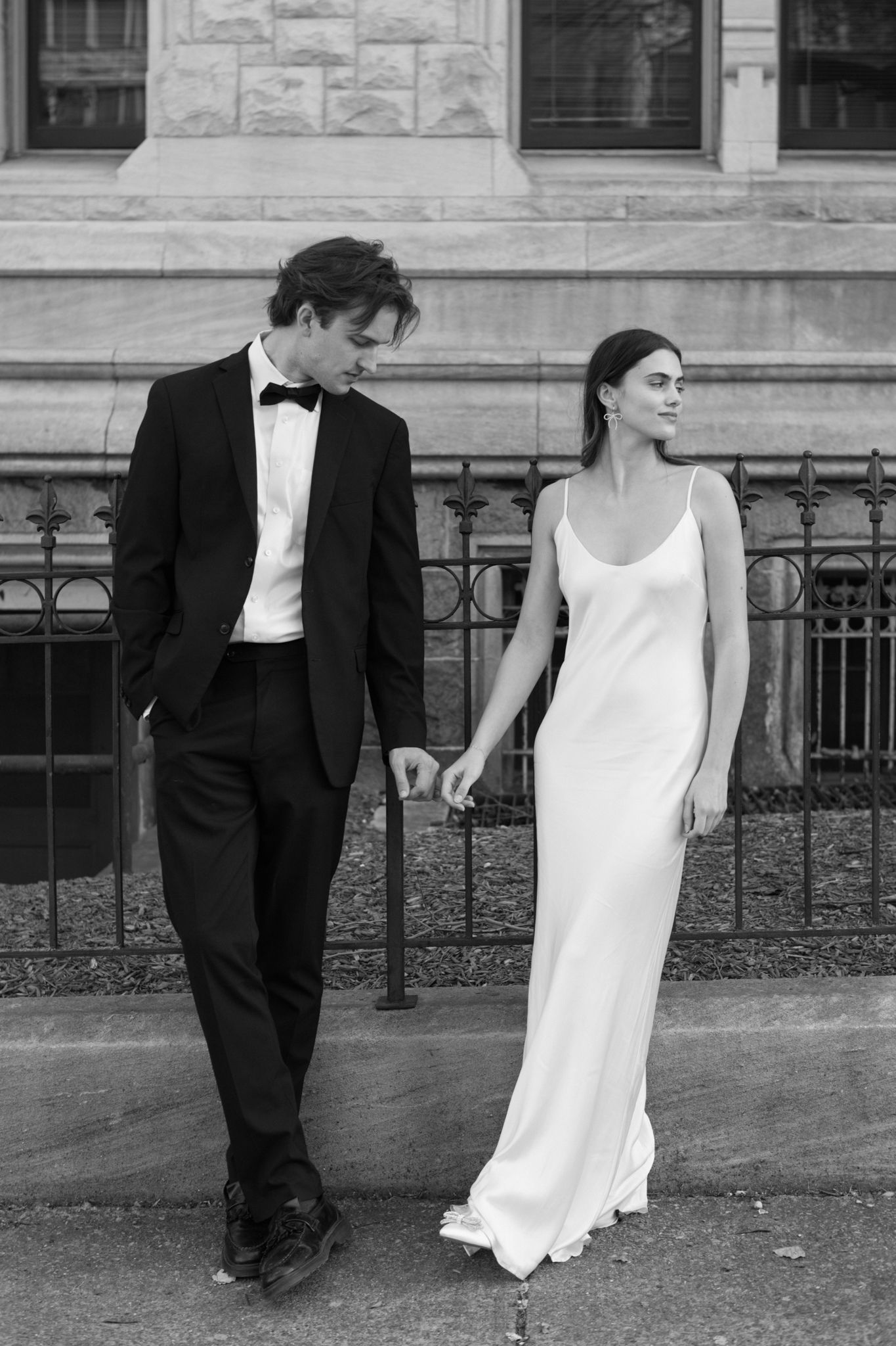 Black and white wedding photo of couple in formal attire standing by iron railing outside stone building.