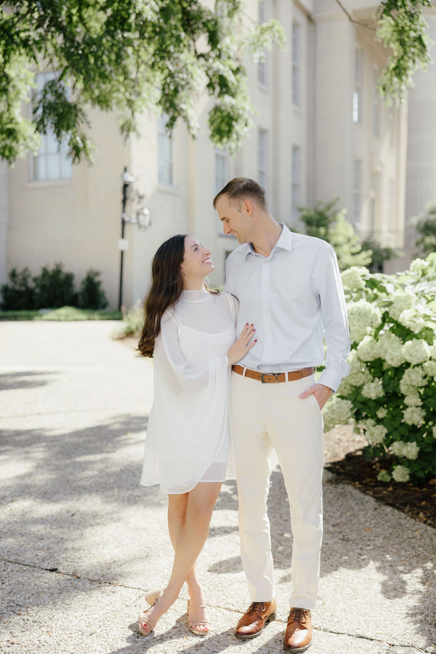 Happy couple in coordinating white outfits gazing at each other near blooming hydrangeas outside a classic building.