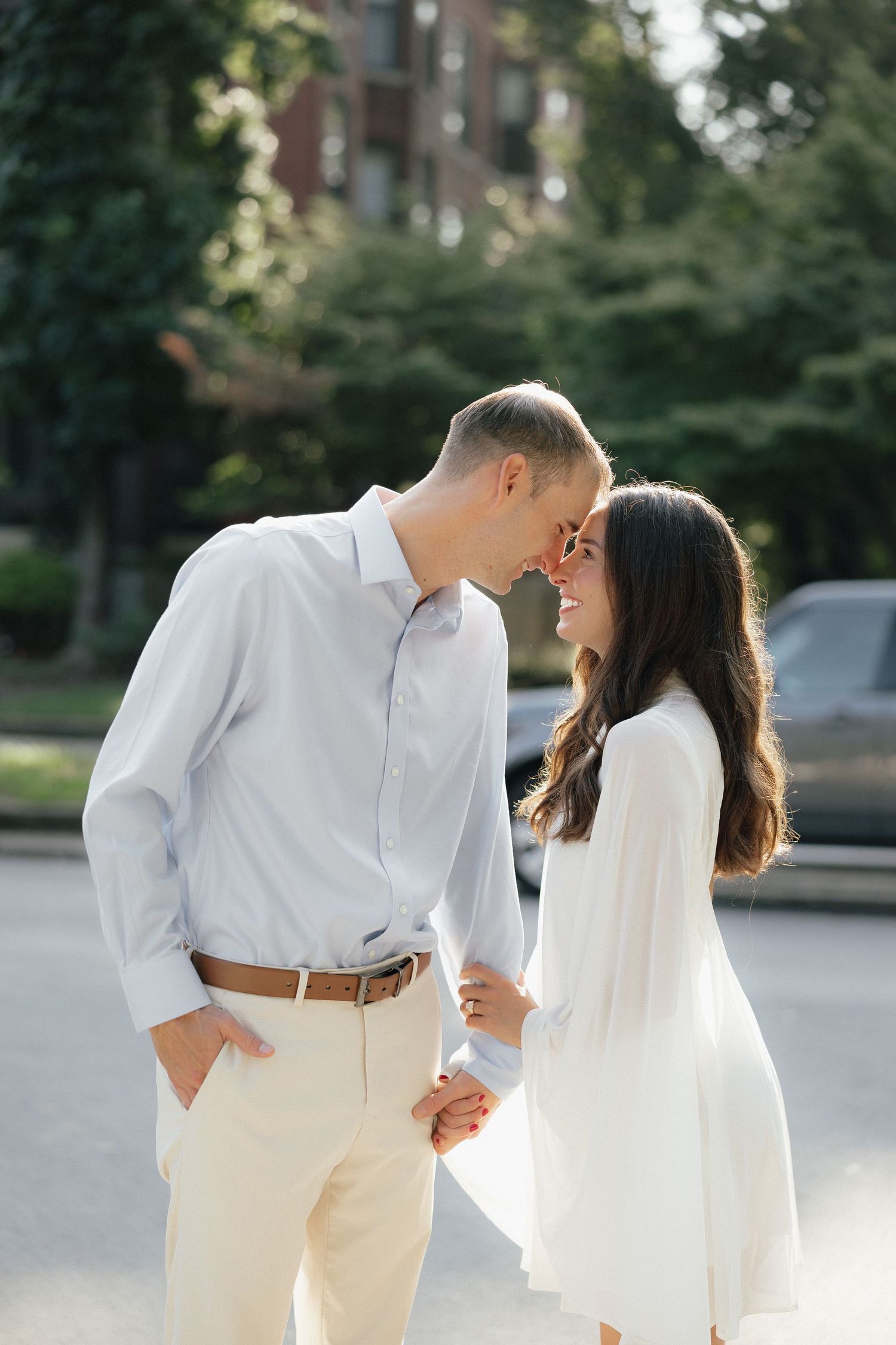 Couple smiling at each other outdoors, man in white shirt and woman in white dress, holding hands on a sunny day.
