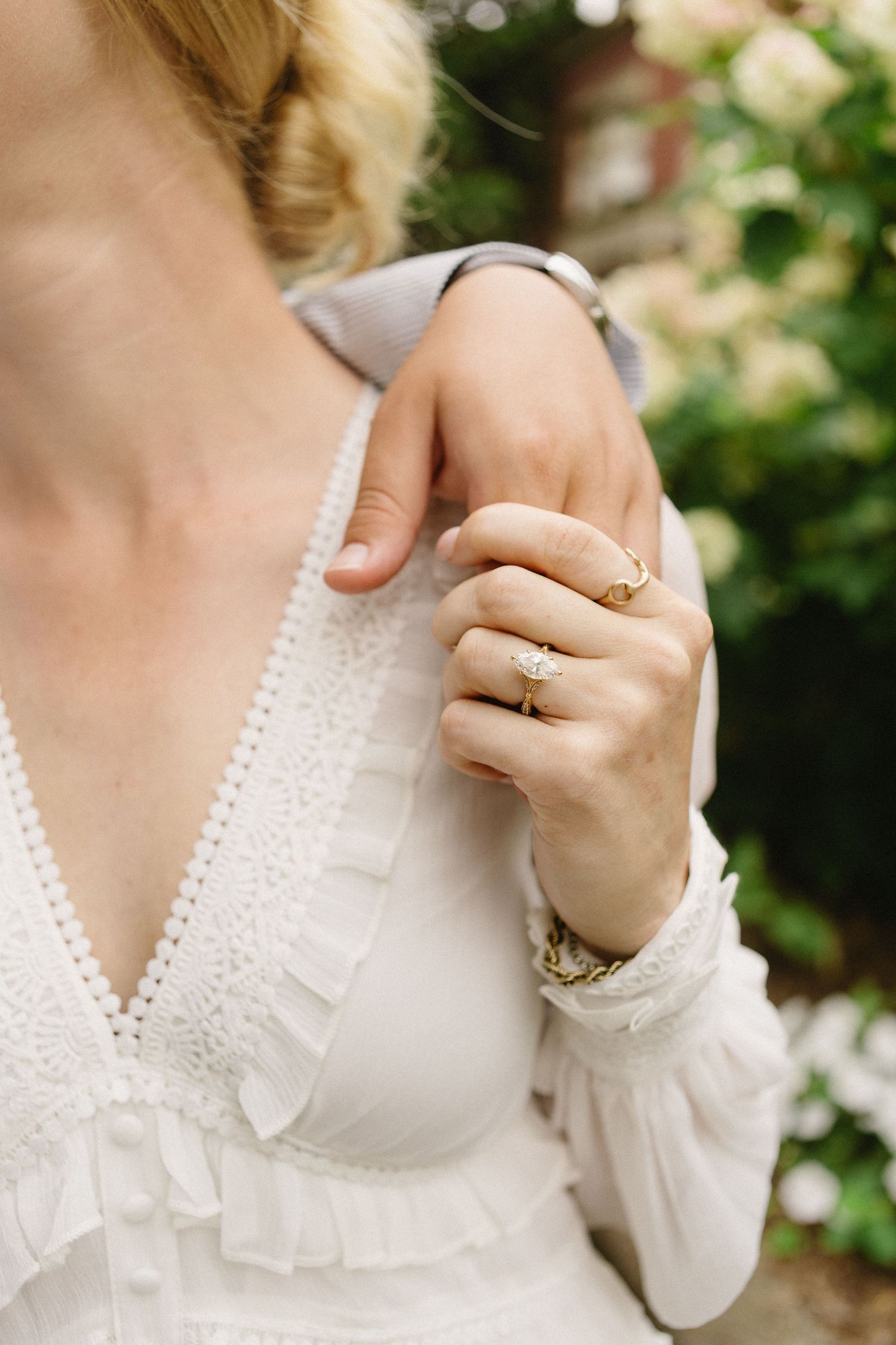Close-up of bride's hand wearing gold engagement ring on white lace wedding dress shoulder.