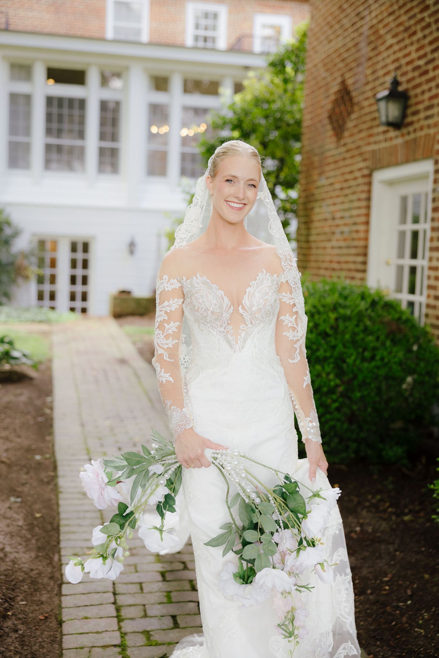 Smiling bride in lace long-sleeve wedding gown holding white floral bouquet outside brick venue.