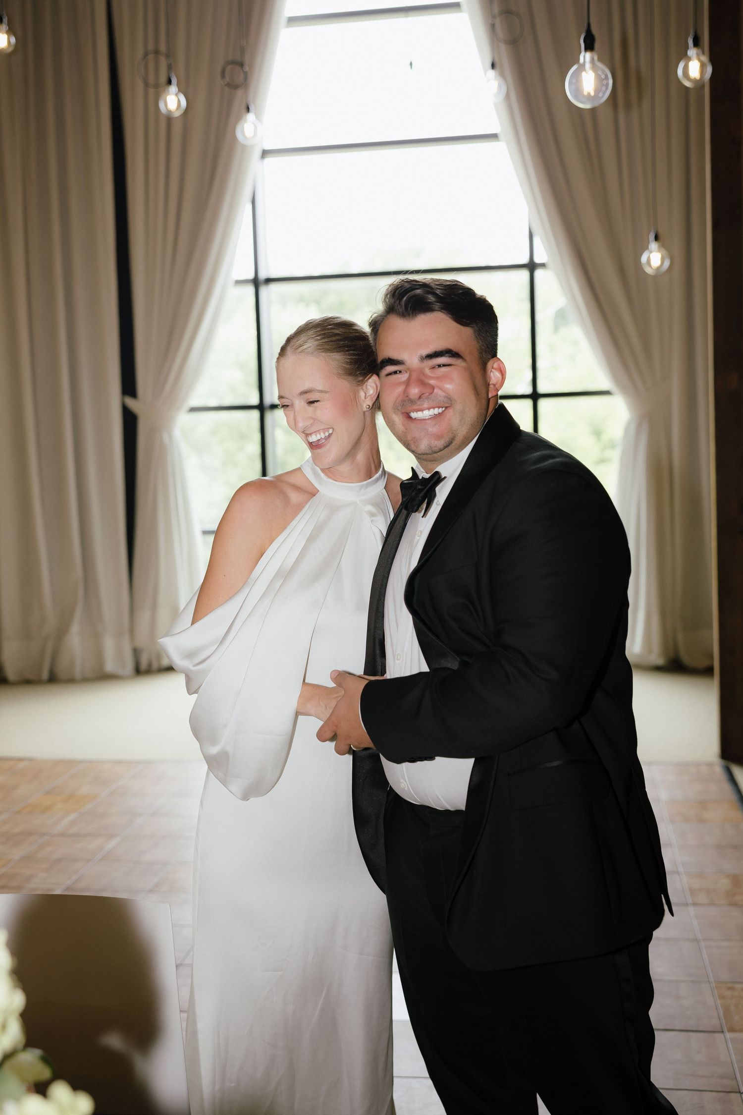 Joyful bride in halter wedding dress and groom in black tuxedo embracing at elegant reception venue.